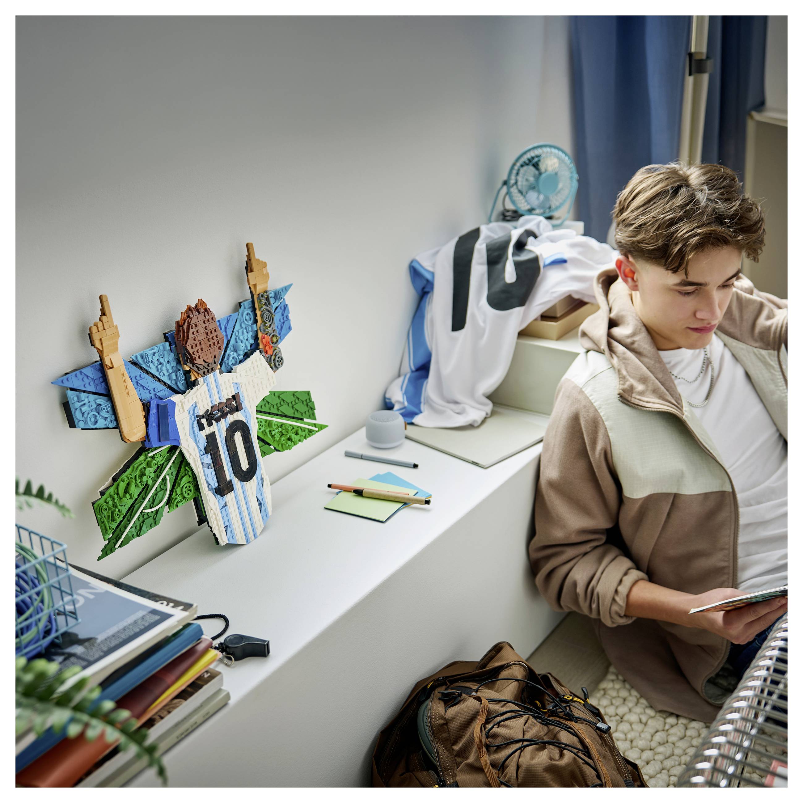 Ein Teenager-Junge sitzt auf dem Boden und liest auf einem Tablet neben einem Schreibtisch mit einem fußball-thematierten Trikot-Bastelwerk und Schulmaterialien in einem Schlafzimmer.