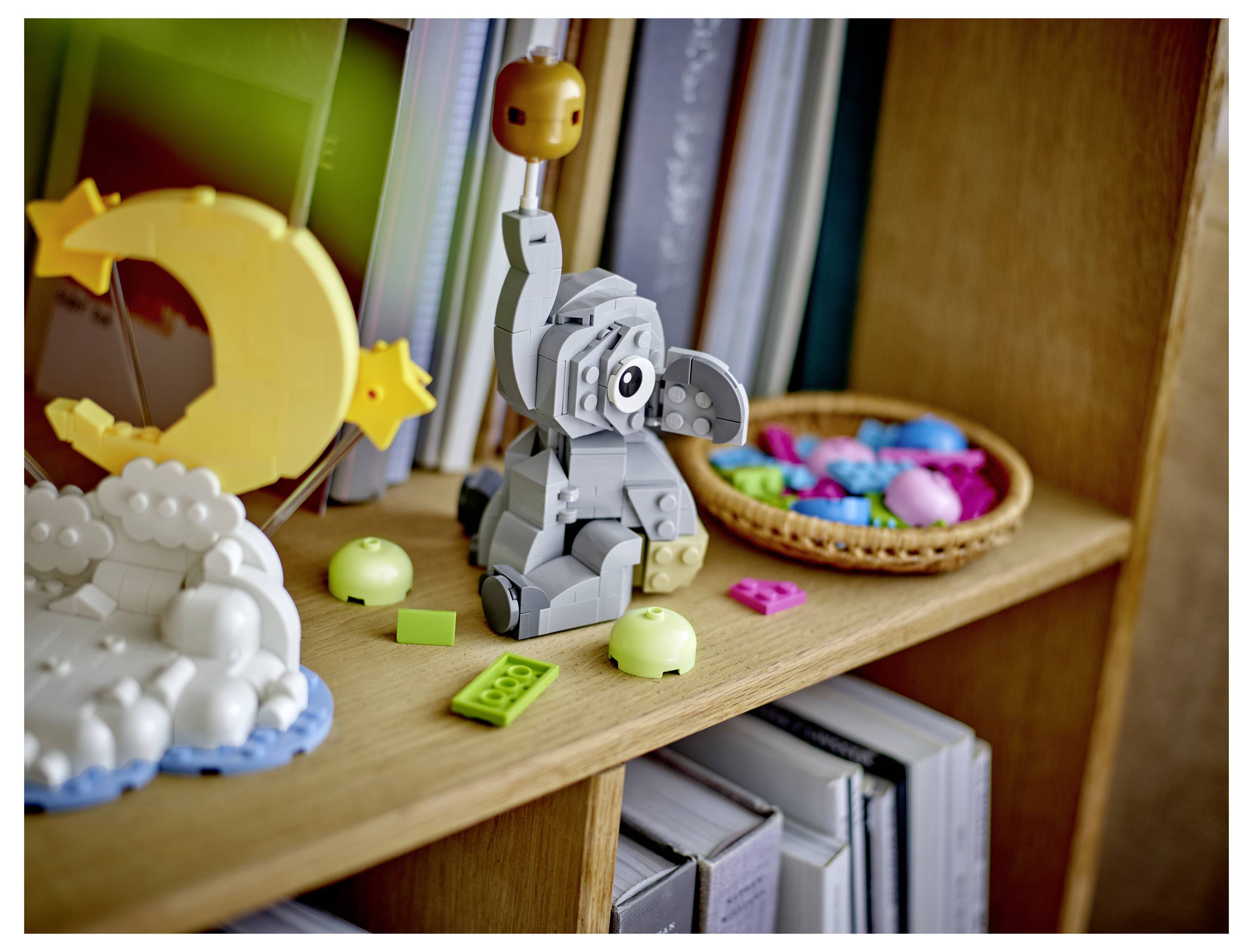 Eine kleine graue Lego-ähnliche Elefantenfigur sitzt auf einem Holzregal neben einer gelben Mondsichel-Dekoration und einem Korb mit bunten Spielzeugteilen.