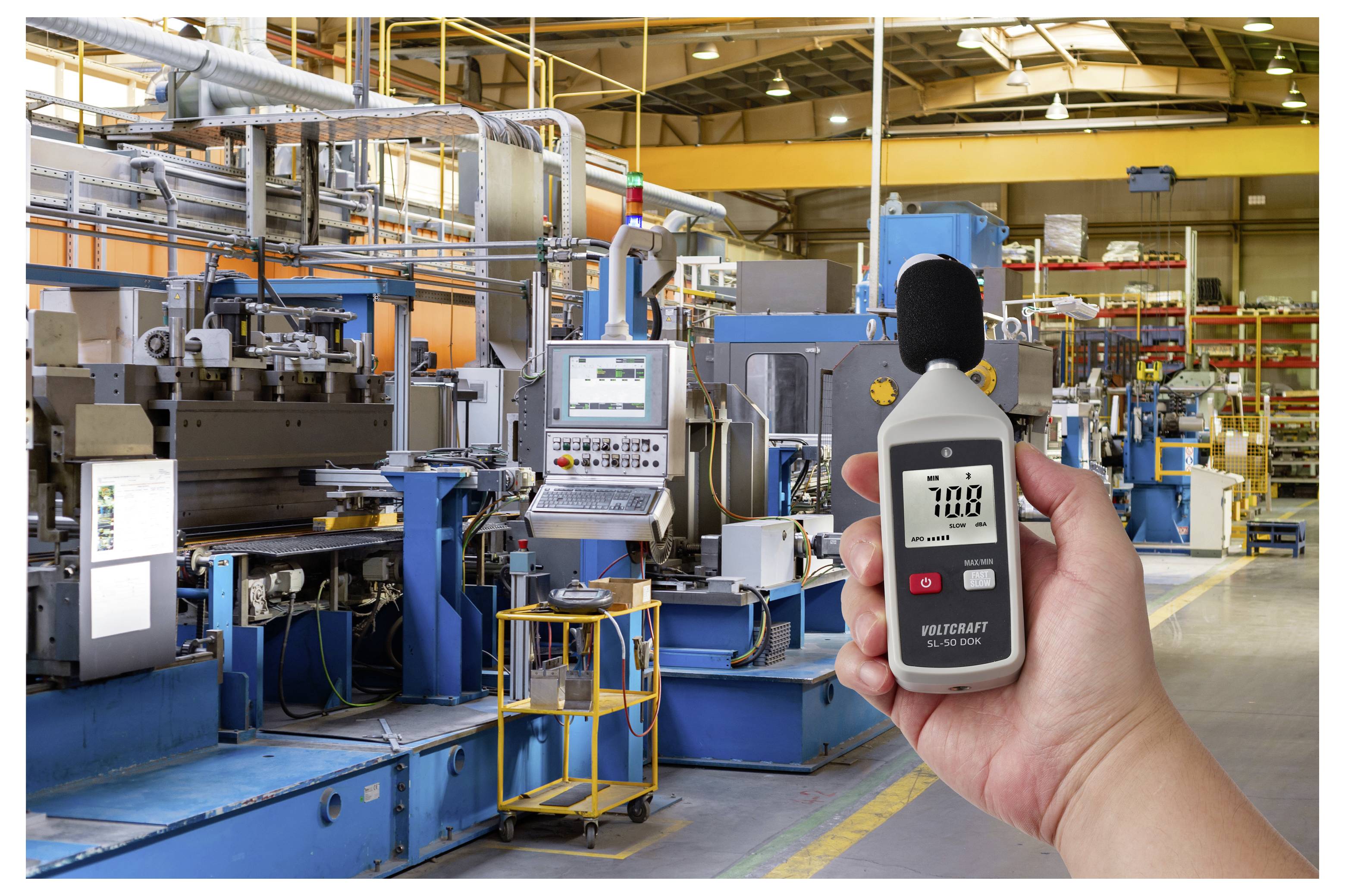 Hand hält ein digitales Schallpegelmessgerät, das 70,8 dBA in einer lauten Fabrik mit großen Industriemaschinen anzeigt.