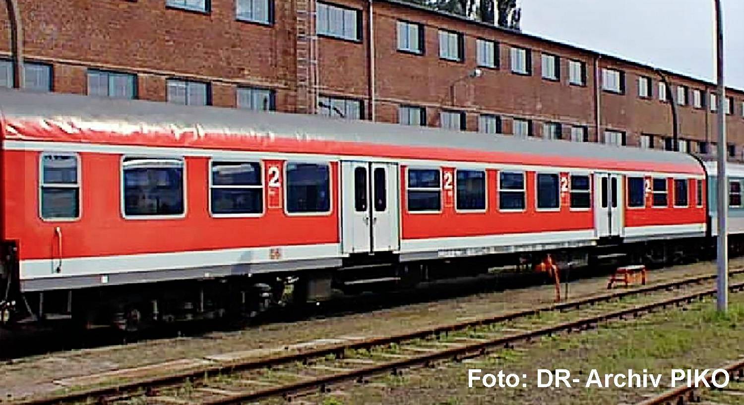 Rot-weiße Fahrgastzugwagen auf Eisenbahngleisen neben einem Backsteingebäude, mit dem Text „Foto: DR-Archiv PIKO