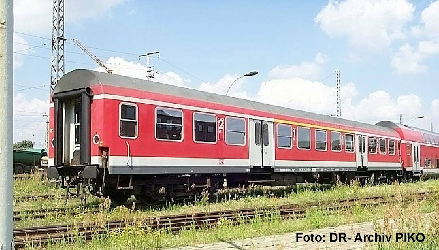 Rot-grauer Personenzugwagen auf Eisenbahnschienen unter blauem Himmel mit einem Wasserzeichen „Foto: DR-Archiv PIKO