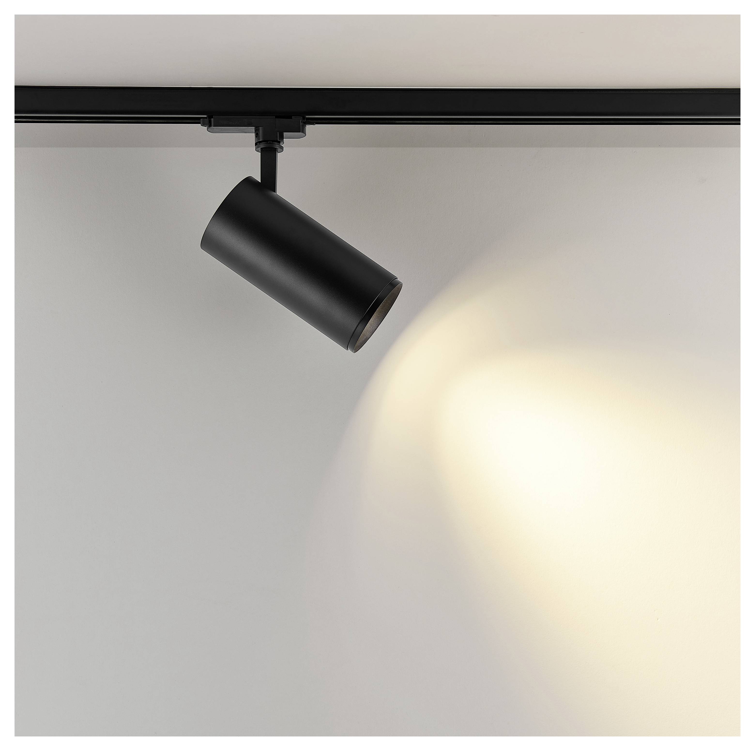 Schwarze Schienenleuchte an der Decke montiert, im Winkel angebracht, um ein warmes Spotlicht auf eine schlichte Wand zu werfen.