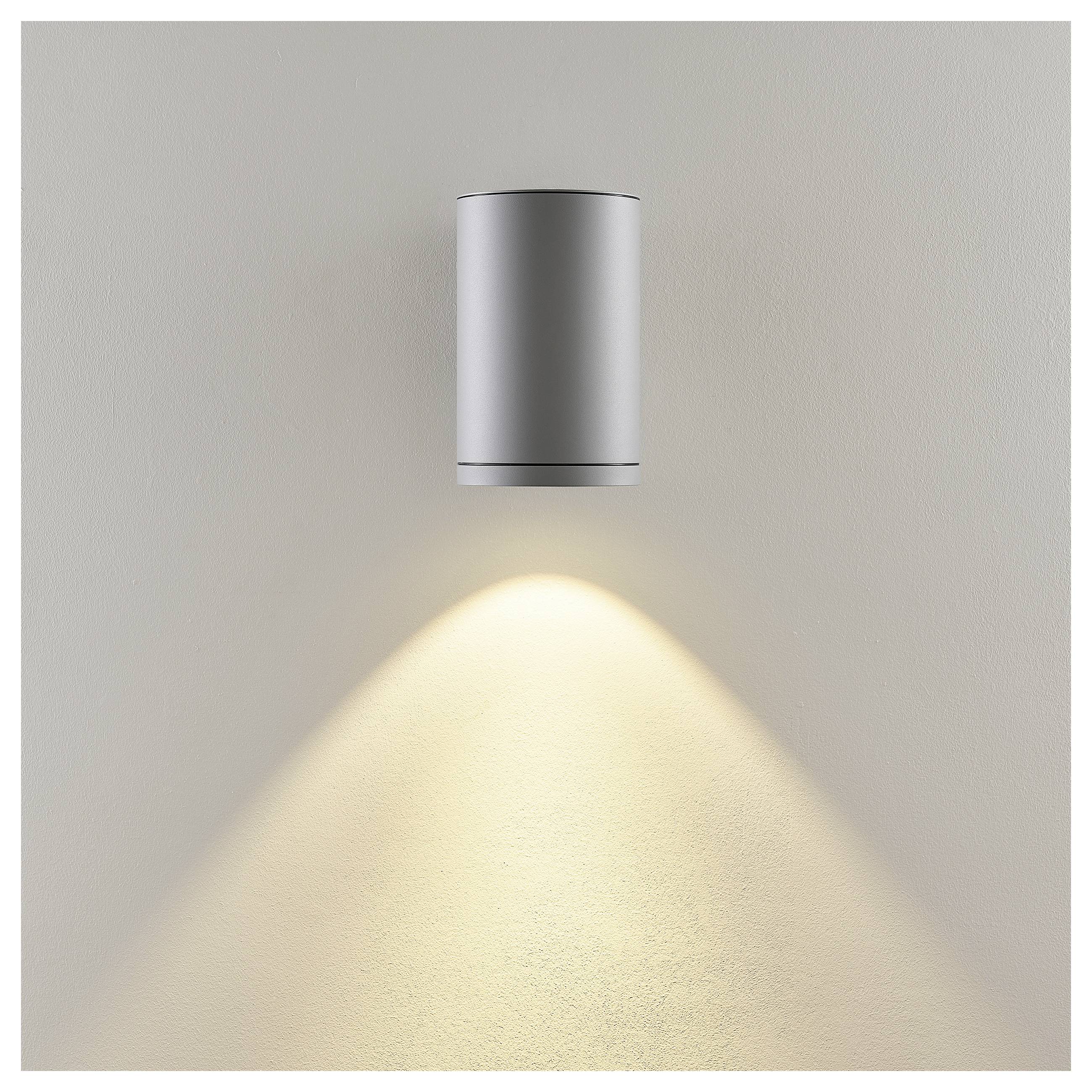 An der Wand montierte silberne zylindrische Leuchte, die einen warmen abwärts gerichteten Lichtkegel auf eine schlichte beige Wand wirft.