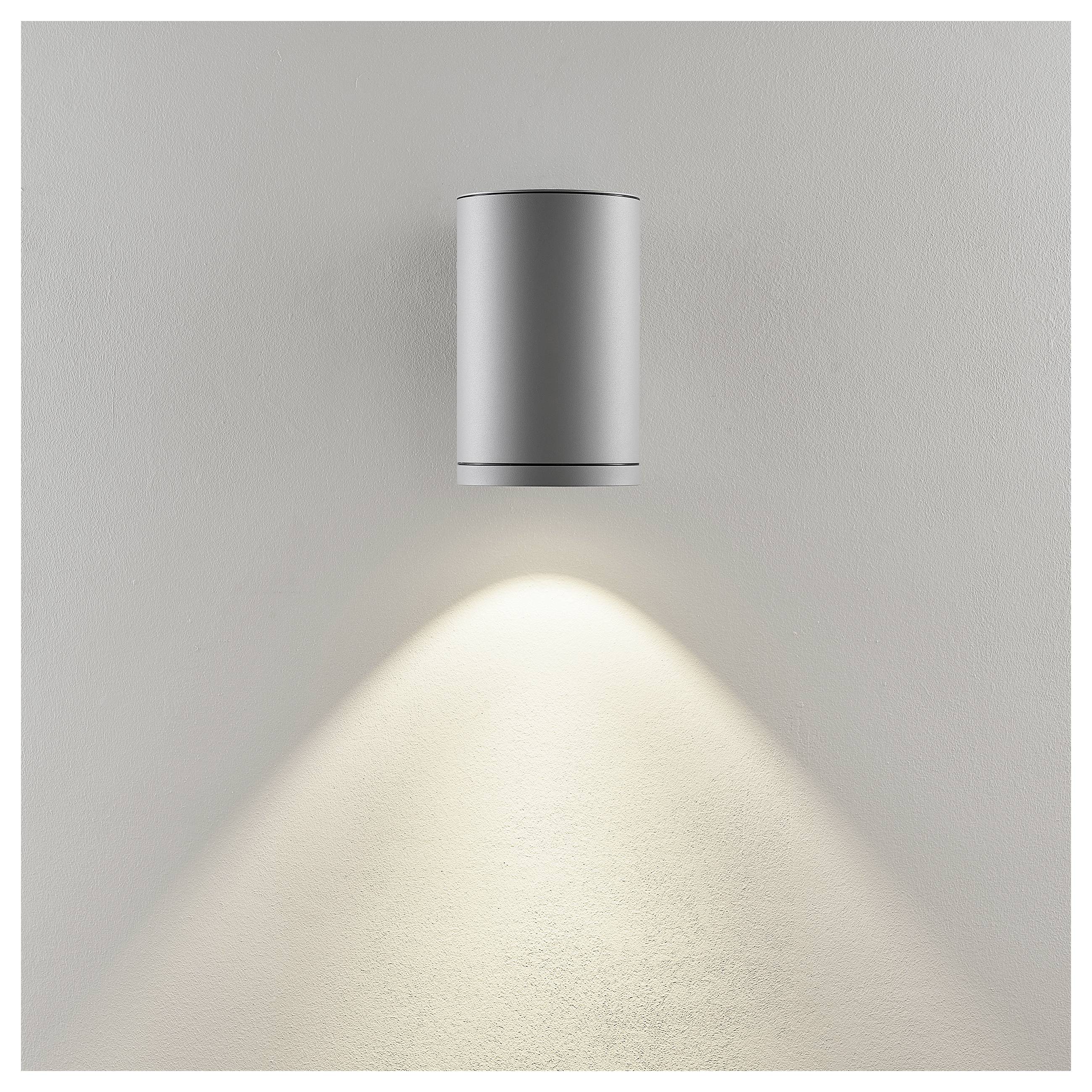 Silberne zylindrische Wandleuchte, die einen warmen Lichtkegel nach unten auf eine schlichte hellgraue Wand wirft.