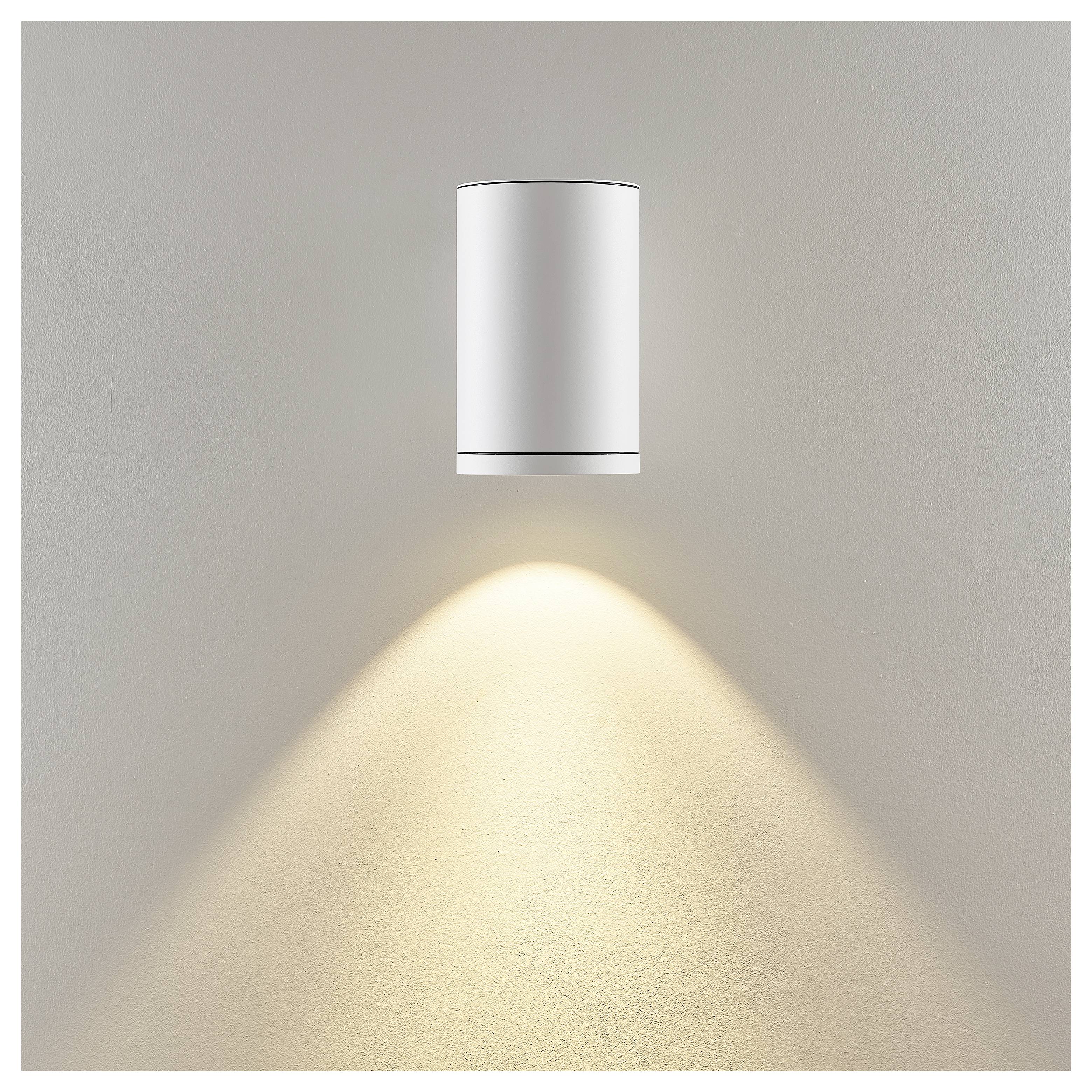Eine minimalistische Wandleuchte mit einem zylindrischen weißen Schirm wirft einen warmen, nach unten gerichteten Lichtkegel auf eine beige Wand.