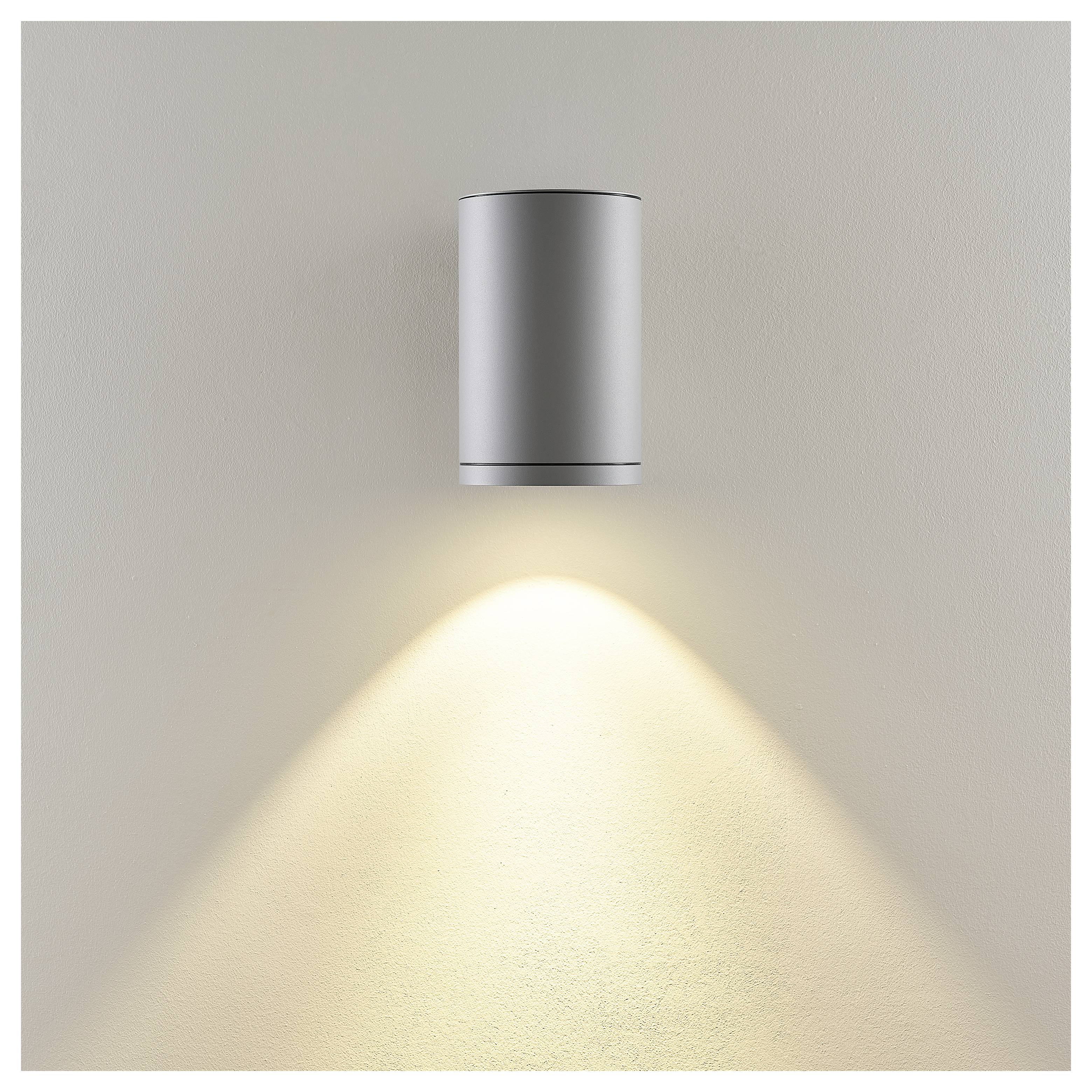 An der Wand montierte silberne zylindrische Leuchte, die einen warmen Lichtkegel auf eine beige Wand wirft.