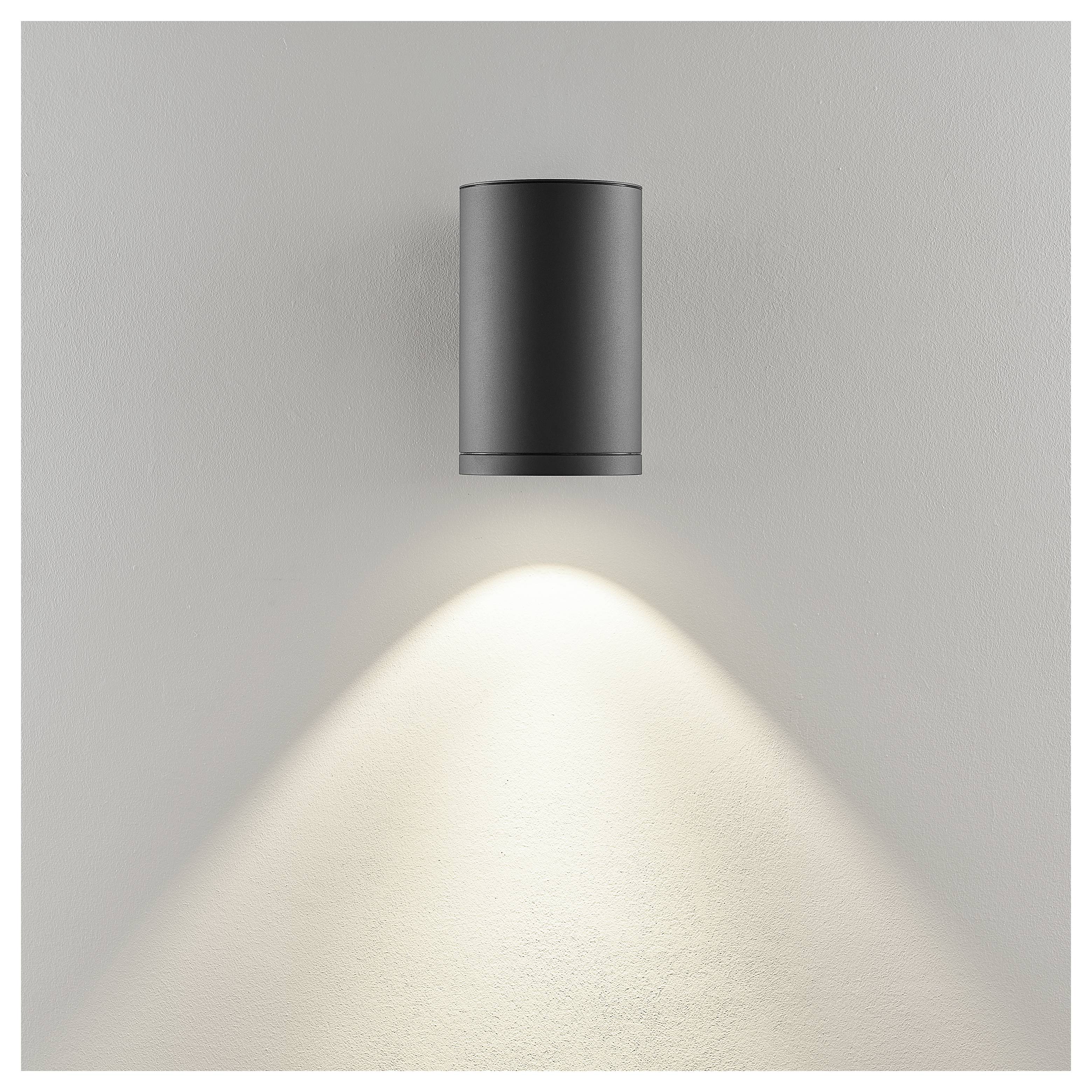 Schwarze zylindrische Wandleuchte an einer grauen Wand, die einen warmen Lichtkegel nach unten wirft.