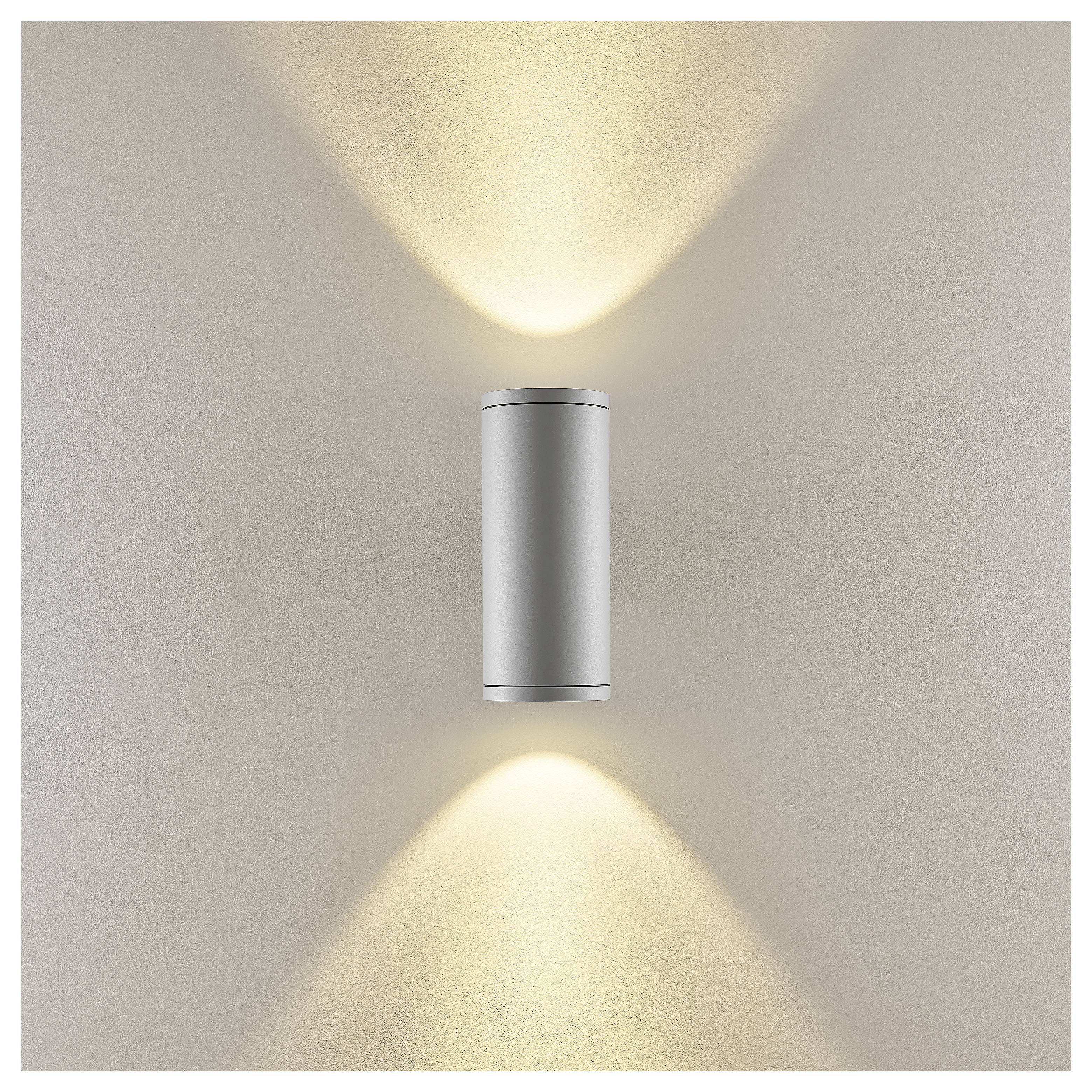 Moderne zylindrische Wandleuchte, die warme Licht­strahlen nach oben und unten auf eine beigefarbene Wand wirft.