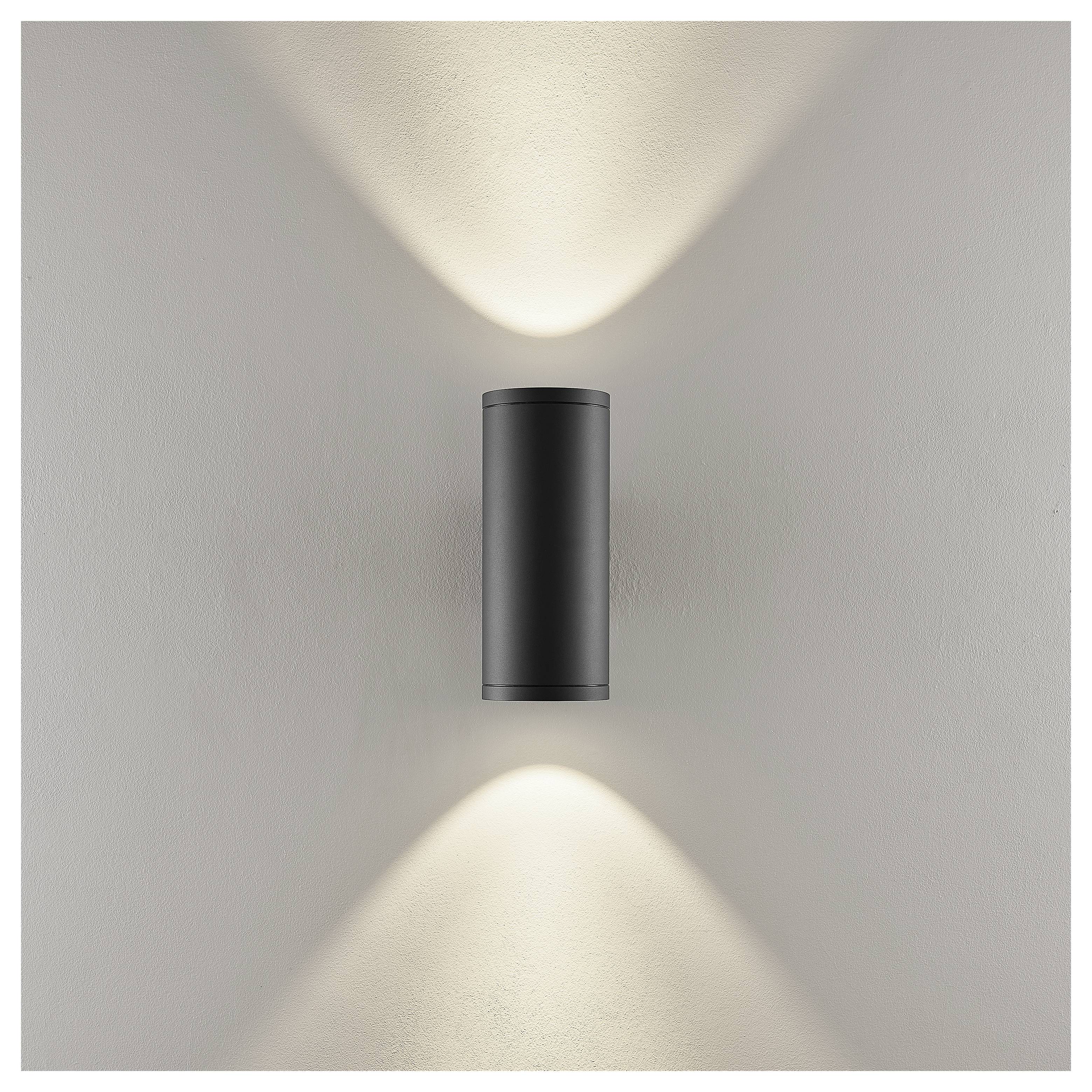 Schwarze zylindrische Wandleuchte, die warme Lichtstrahlen nach oben und unten auf eine hellgraue Wand wirft.