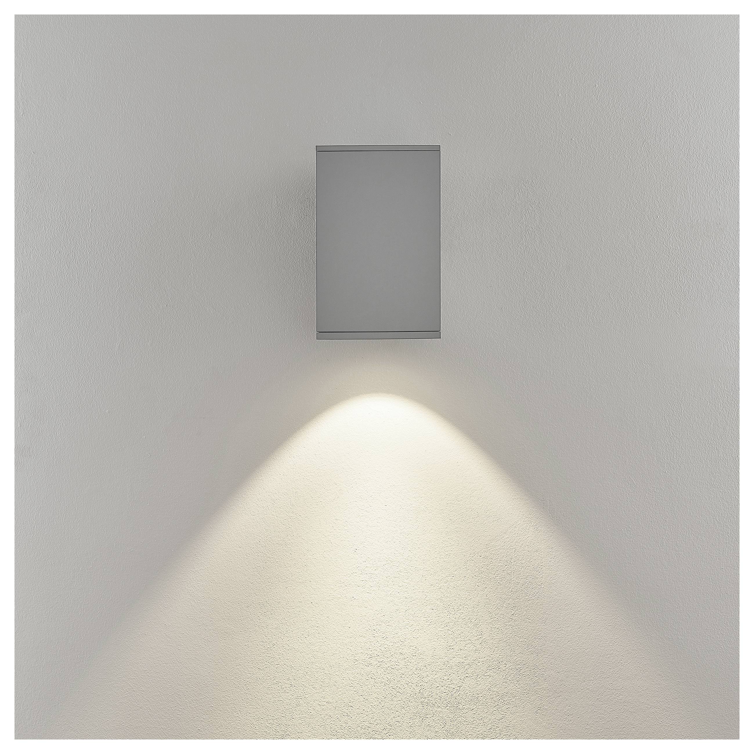 Minimalistische Wandleuchte mit rechteckiger grauer Abdeckung wirft einen weichen, nach unten gerichteten Lichtkegel auf eine weiße Wand.