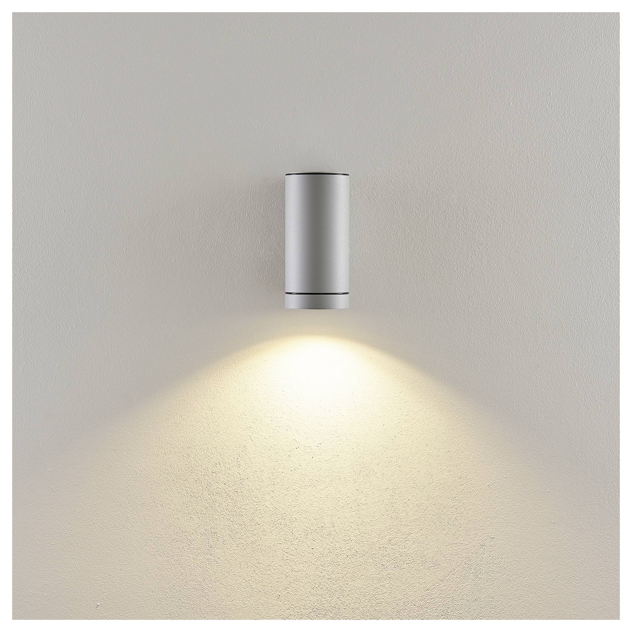 An der Wand montierte zylindrische Leuchte, die einen warmen Lichtkegel nach unten auf eine schlichte beige Wand wirft.