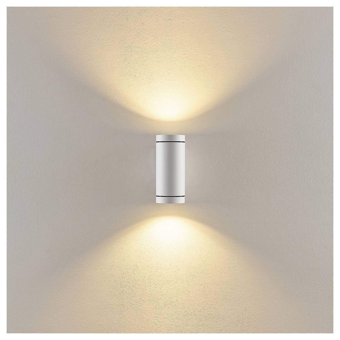An der Wand montiertes zylindrisches Leuchtenelement, das warme Lichtstrahlen nach oben und unten auf eine beige Wand wirft.