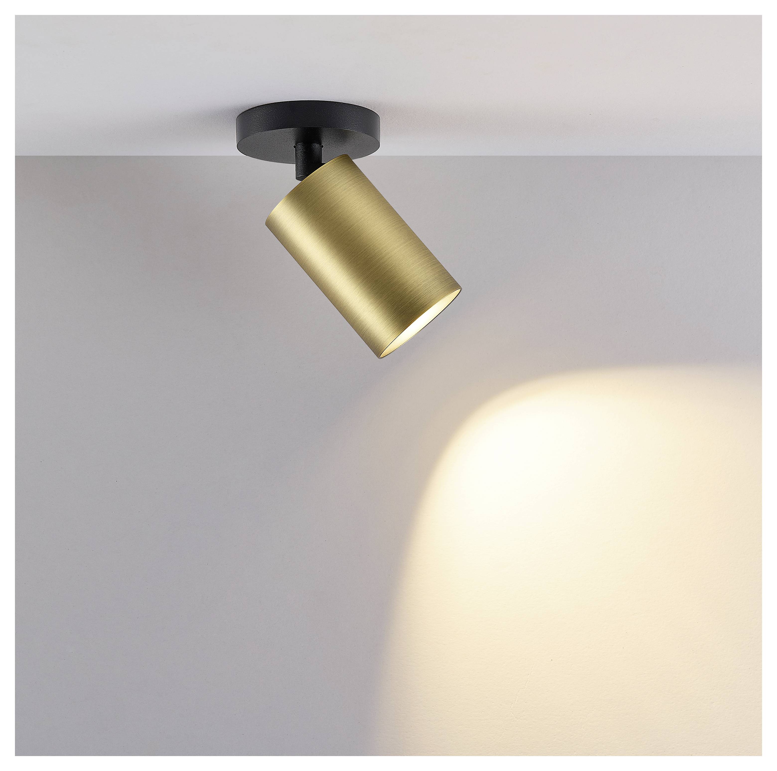 Goldener zylindrischer Scheinwerfer an der Decke montiert, der einen warmen Strahl auf eine schlichte graue Wand wirft.