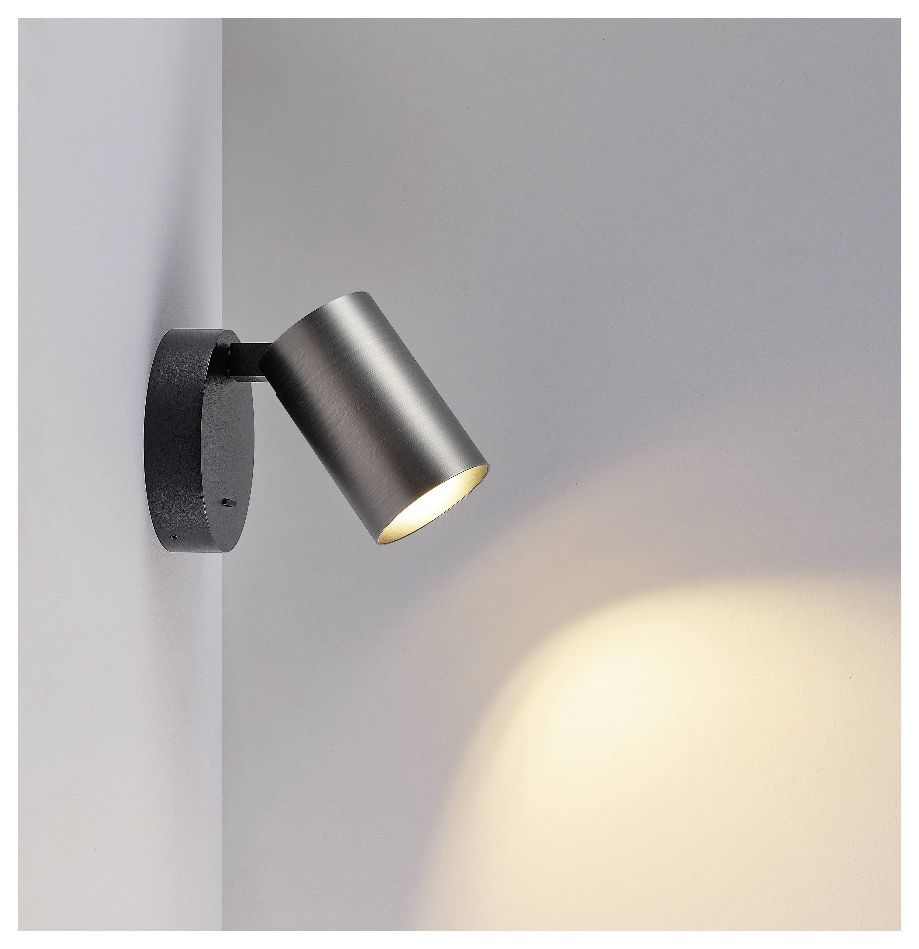 Moderner wandmontierter Strahler mit schwarzer Basis und gebürstetem Metallschirm, der einen warmen Lichtstrahl auf eine graue Wand wirft.