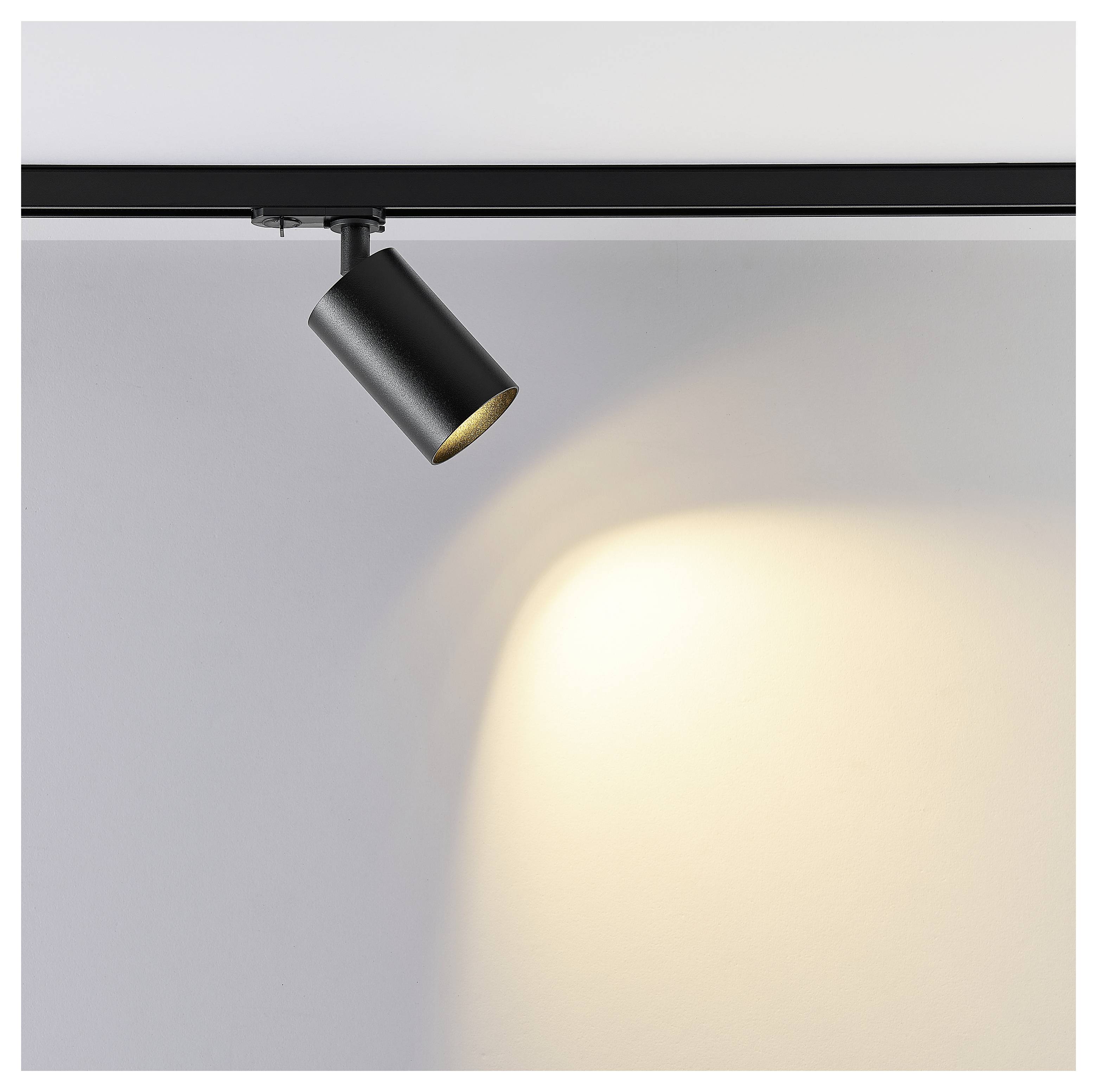 Schwarzer Strahler an einer Deckenleiste montiert, der einen warmen Lichtkegel auf eine Wand wirft.