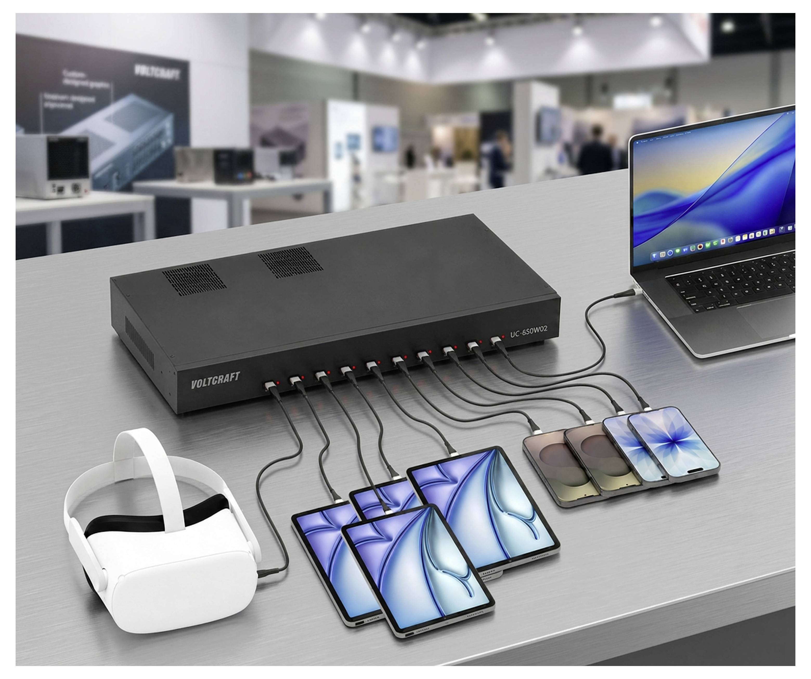 Ein USB-Ladehub auf einem Tisch versorgt mehrere Smartphones, ein VR-Headset und einen Laptop in einer Messeausstellung mit Strom.