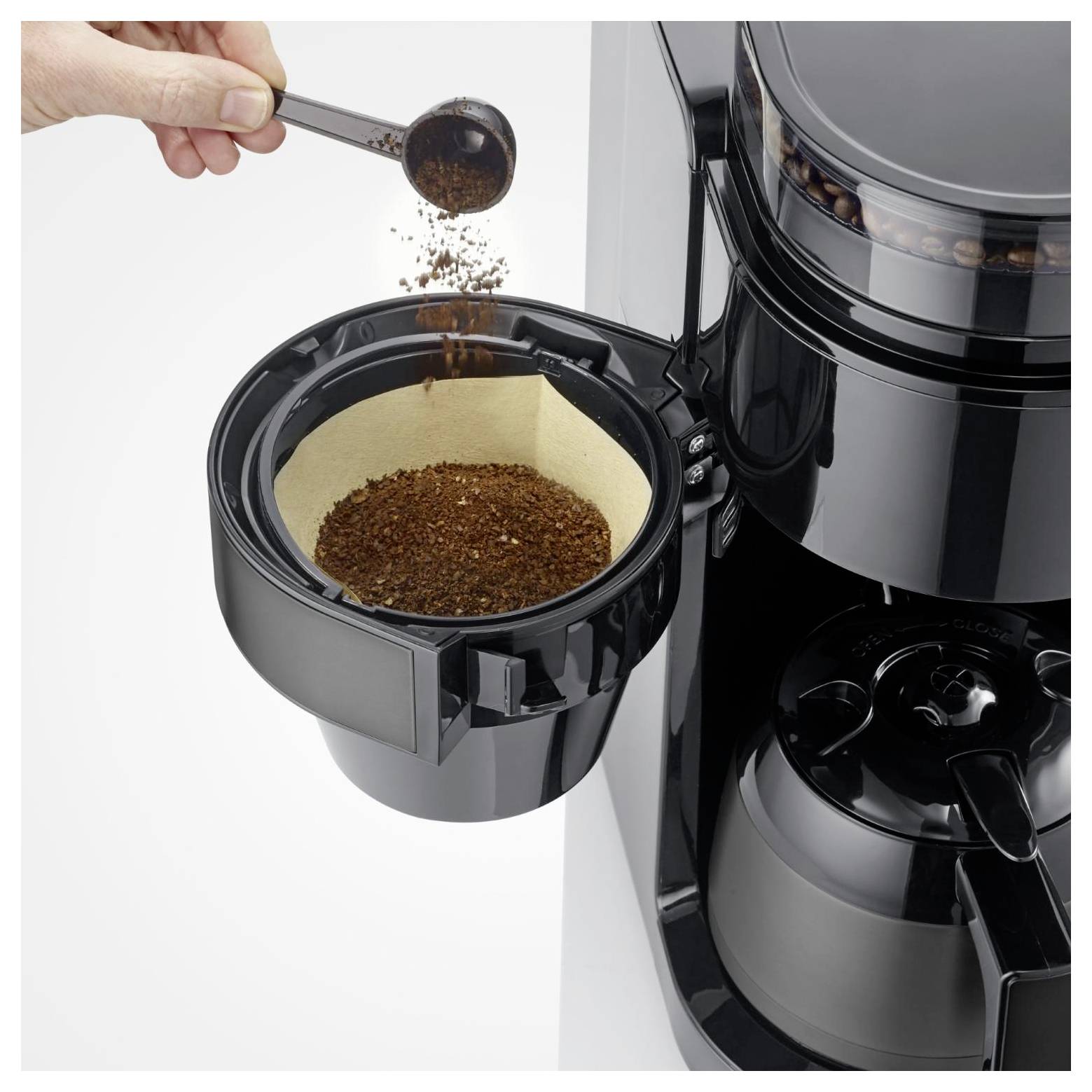 Hand gießt gemahlenen Kaffee in einen abnehmbaren Korb eines schwarzen Tropfkaffeeautomaten.