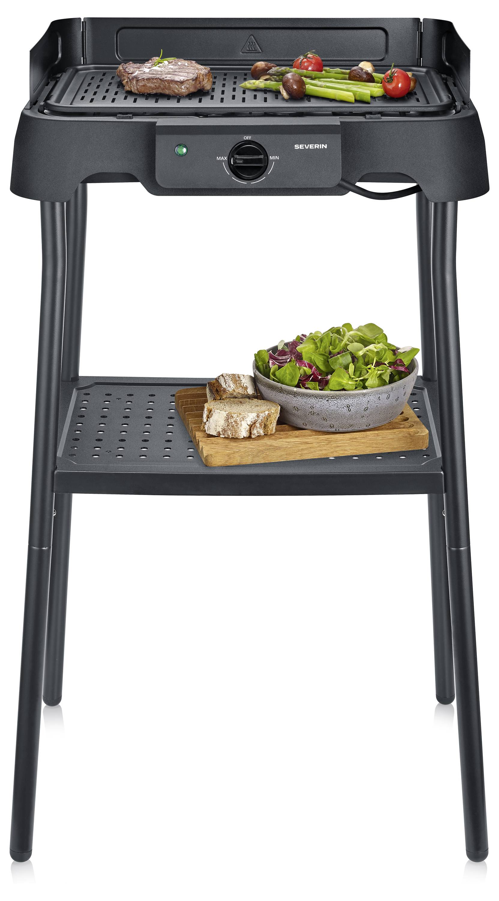 Schwarzer elektrischer Grill mit zwei Regalen, auf dem oben Steak und Gemüse kochen und auf dem unteren Regal eine Schüssel mit Salat und Brot zu sehen sind.