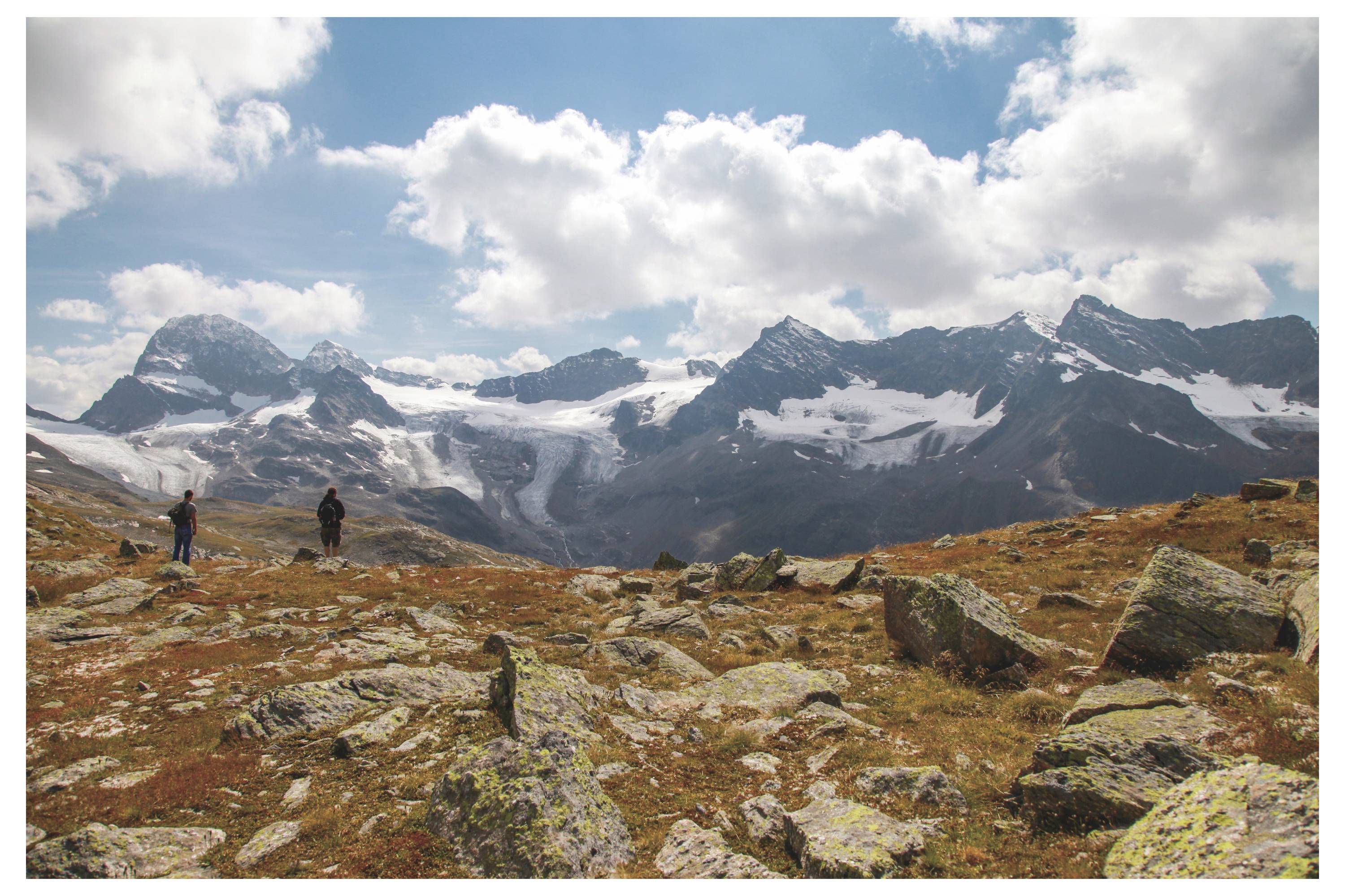 Wanderer auf einem felsigen Alpenweg mit verschneiten Bergen und Gletschern unter bewölktem Himmel.