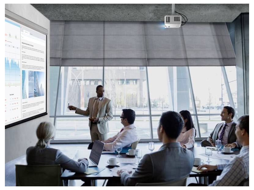 Geschäftspräsentator spricht zu sitzenden Kollegen in einem Konferenzraum und zeigt auf eine Wandanzeige mit Diagrammen und Bildern während einer Besprechung.
