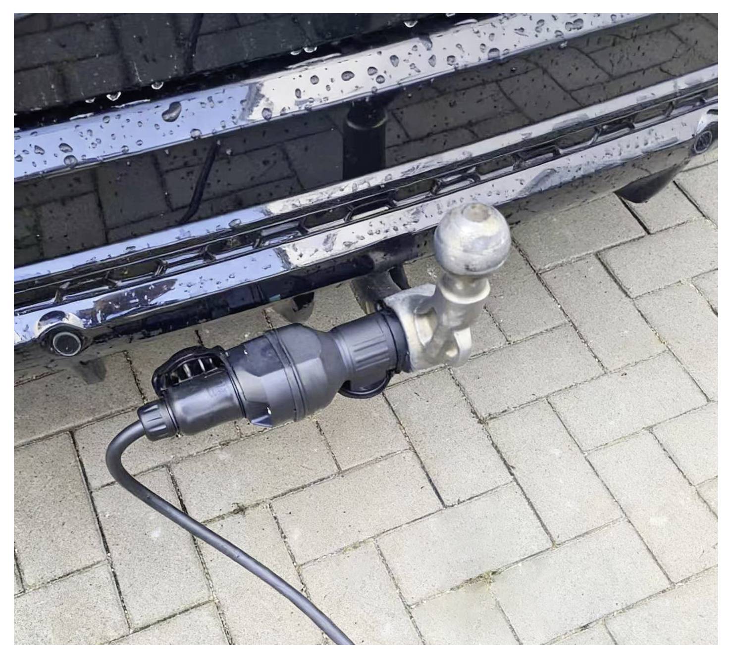 Anhängerkupplung mit angeschlossenem Elektrikadapter in eine Fahrzeugkupplung eingesteckt, in Nahaufnahme auf nassen Pflastersteinen dargestellt.