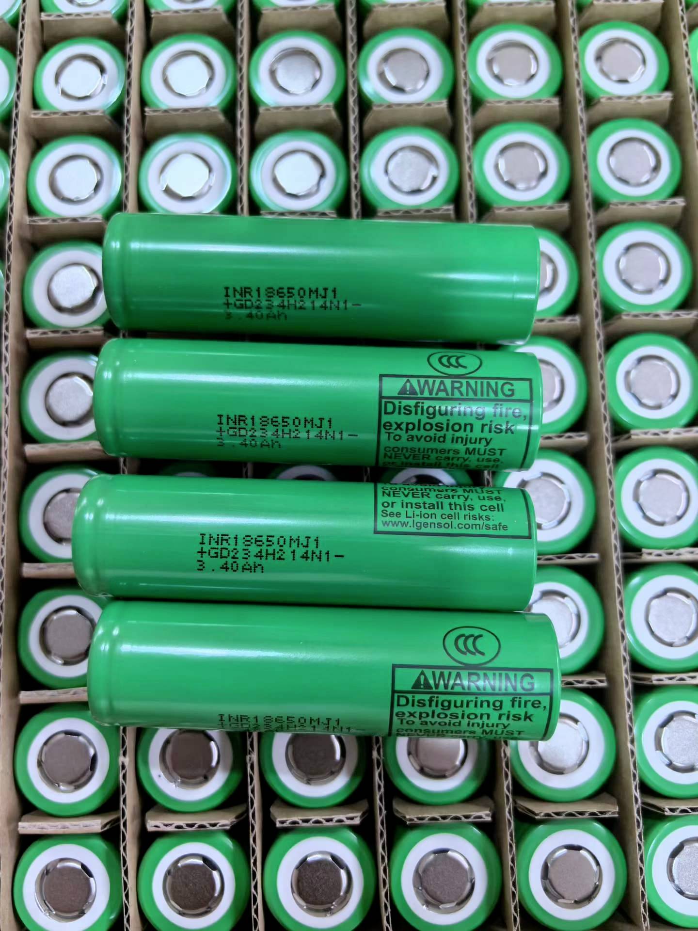 Gestapelte grüne Lithium-Ionen-Batterien in Pappschachteln, mit mehreren größeren Zellen, die mit Warntext über Feuer- und Explosionsgefahr gekennzeichnet sind.