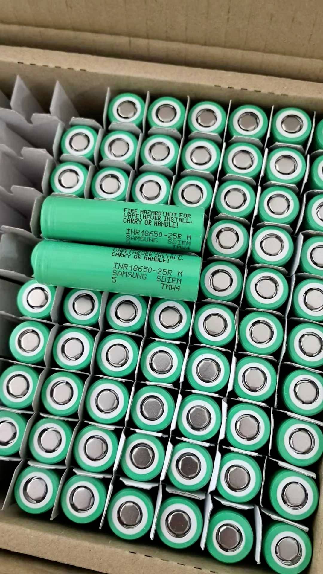 Karton mit grünen zylindrischen Batteriezellen, wobei einige Zellen beschriftet sind und über der Oberseite angeordnet liegen.