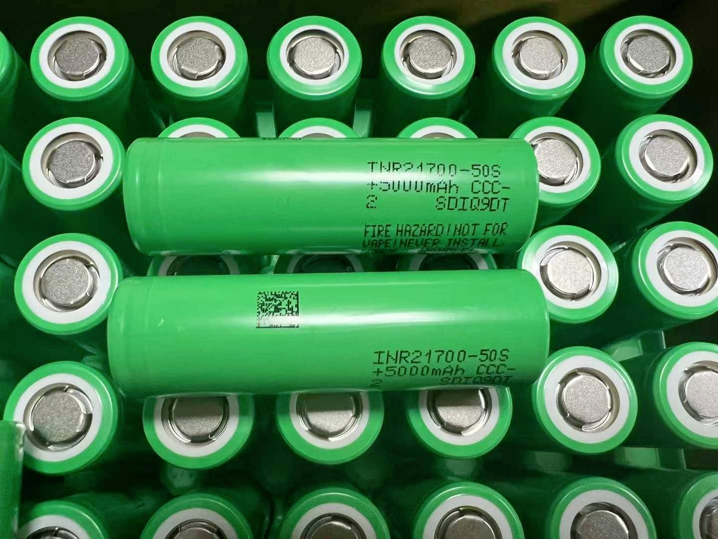Grüne zylindrische Batterien in Reihen gestapelt, mit zwei beschrifteten Zellen, die oben quer angeordnet sind und auf Speicherung oder Überprüfung von wiederaufladbaren Batterien hindeuten.