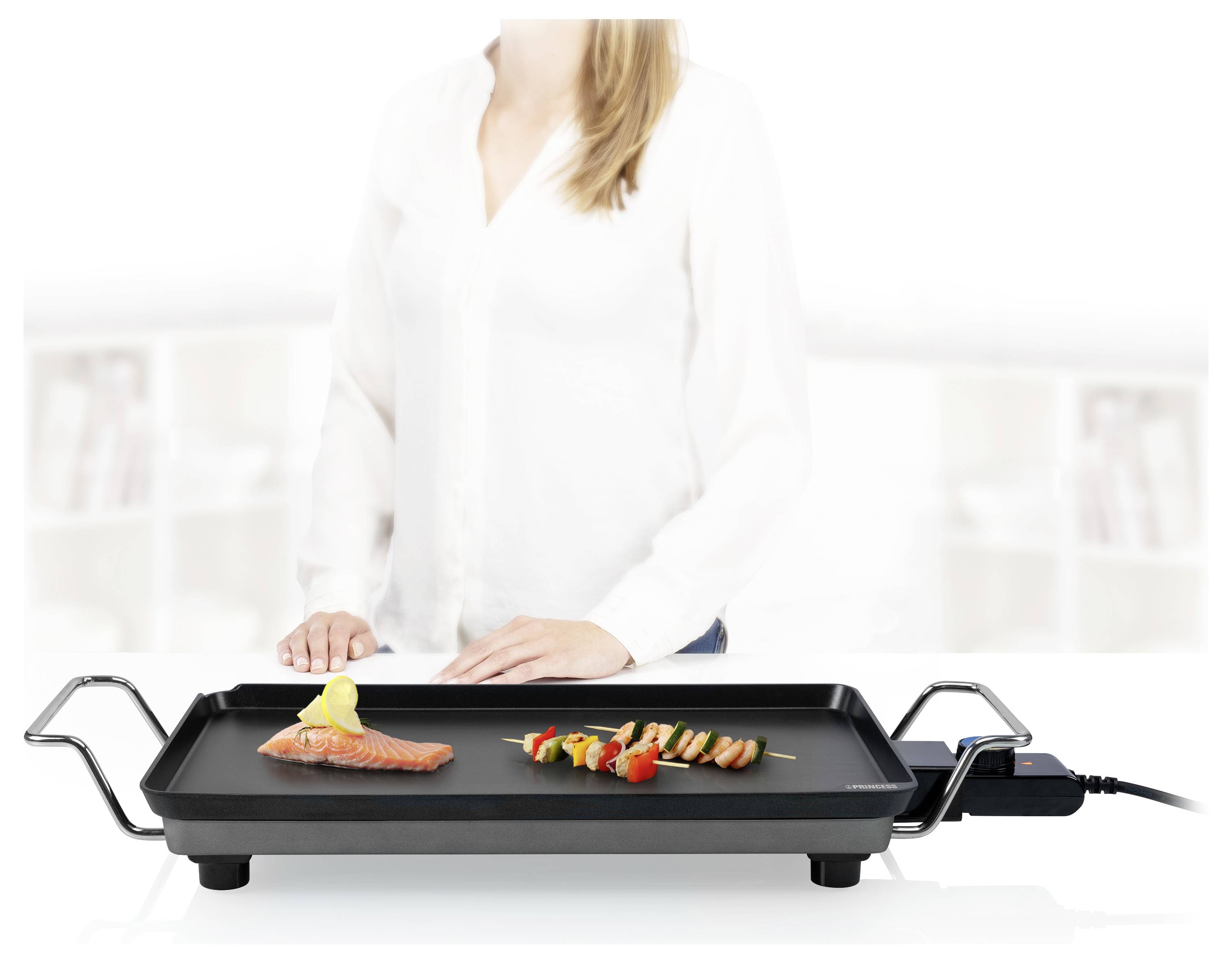 Eine Person bereitet Lachs- und Gemüsespieße auf einer elektrischen Grillplatte in einer hellen Küche zu.