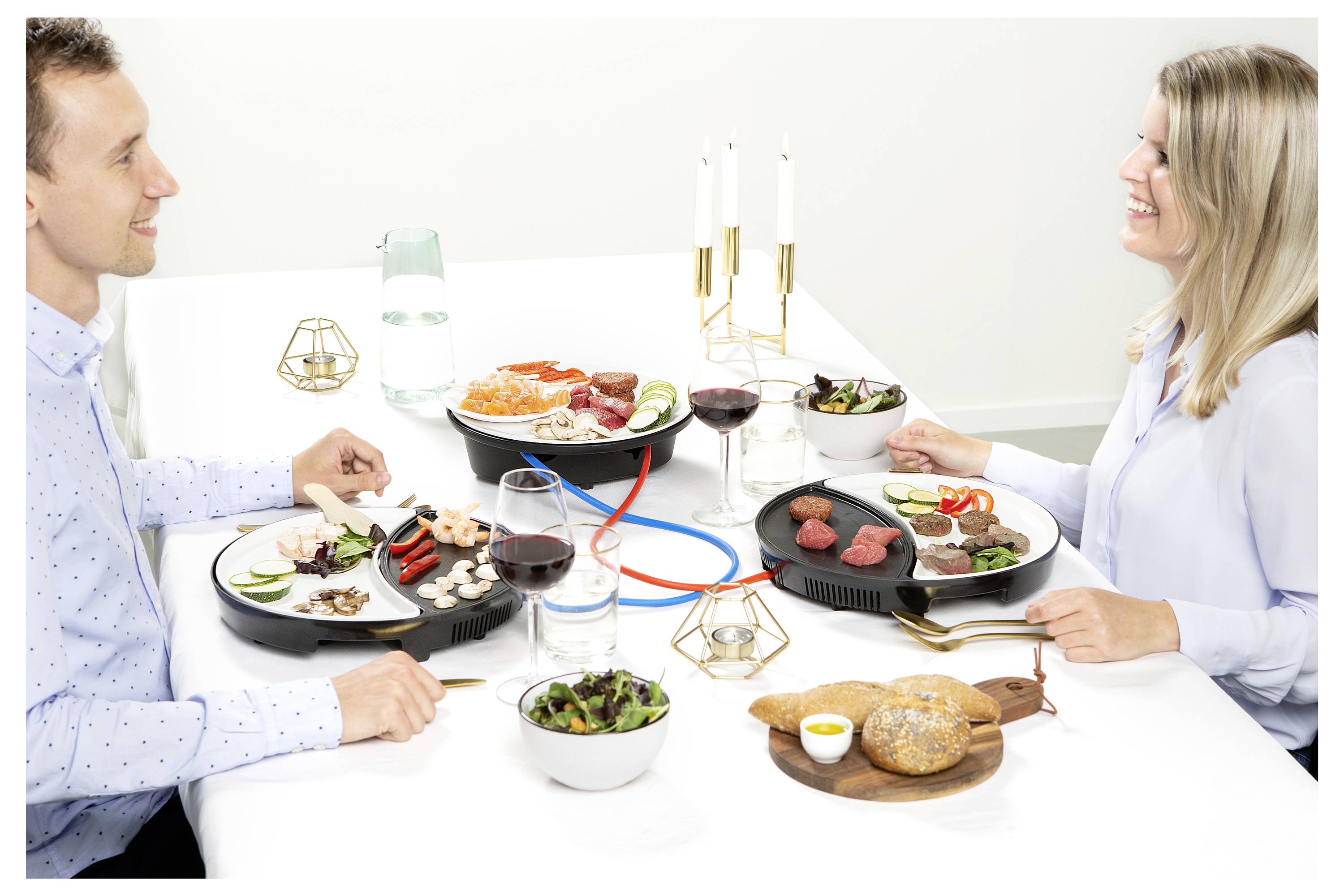 Ein Mann und eine Frau teilen sich ein Fondue-Essen an einem Esstisch mit Tellern voller Fleisch, Gemüse, Brot, Wein und Kerzen.