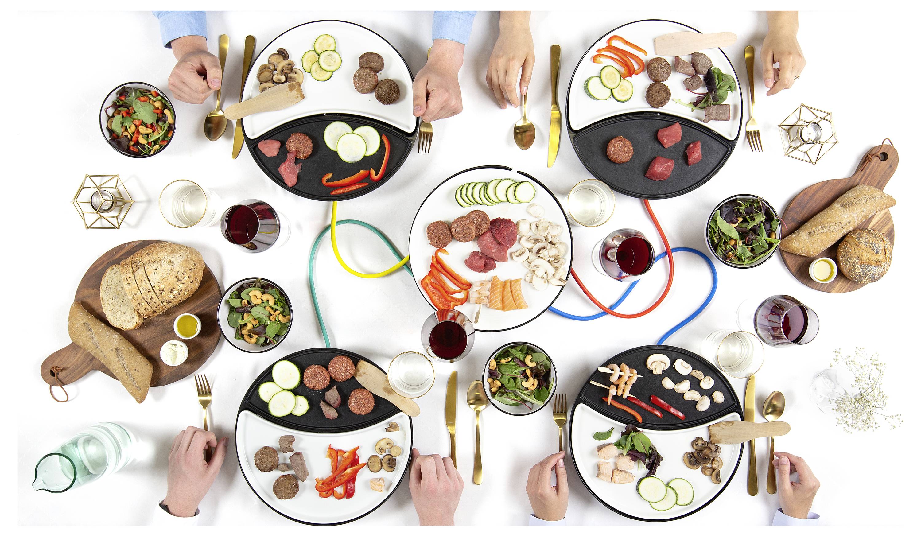 Draufsicht auf sechs Personen, die ein gemeinsames Mahl teilen, mit verschiedenen Brotsorten, Fleisch, Gemüse, Salaten und Getränken auf einem weißen Tisch mit farbigen Kabeln, die die Gedeck verbinden.
