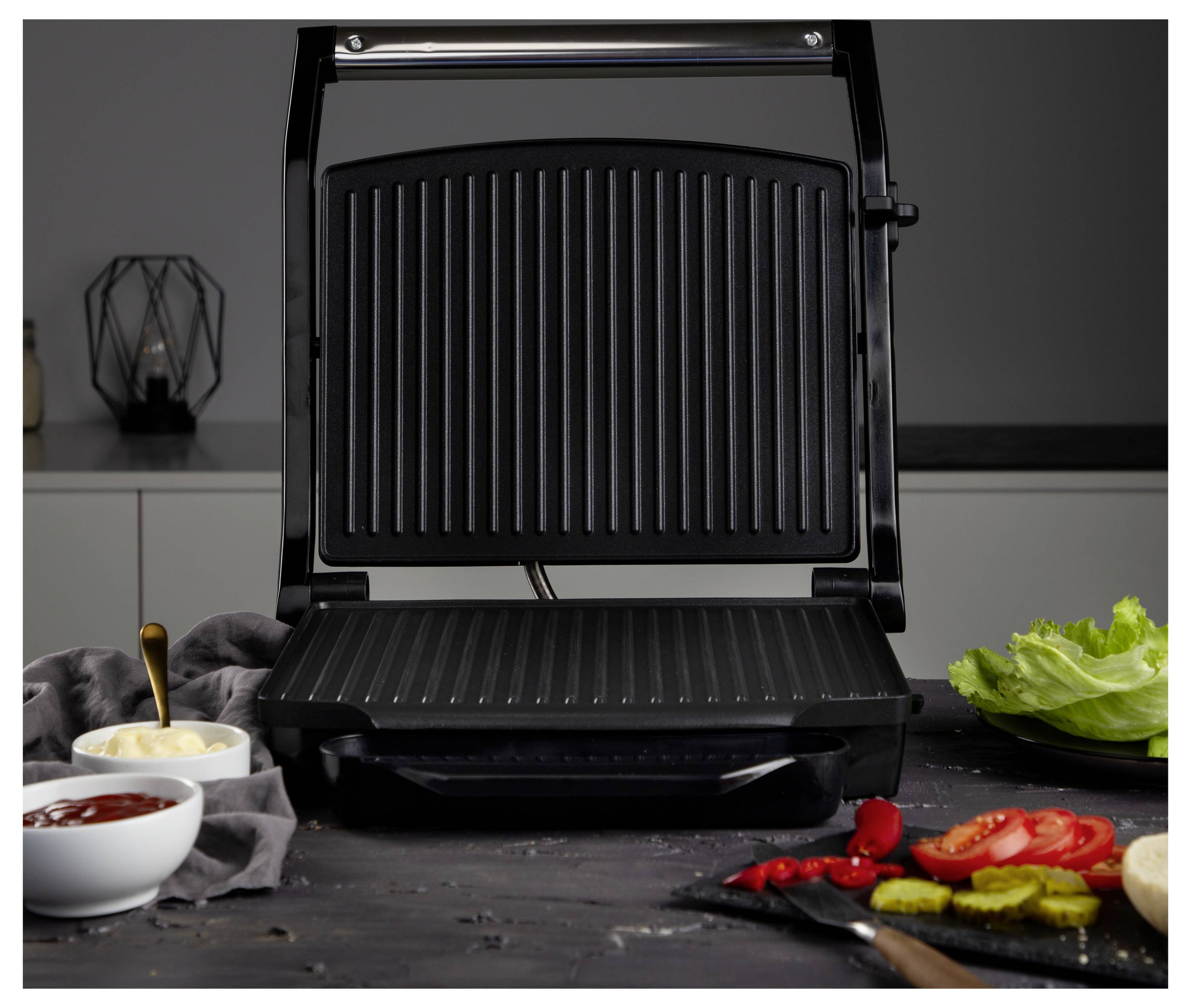Ein offener Tischgrill in der Mitte einer Küchenarbeitsplatte, mit Schüsseln mit Sauce, Salat und geschnittenem Gemüse rundherum angeordnet.