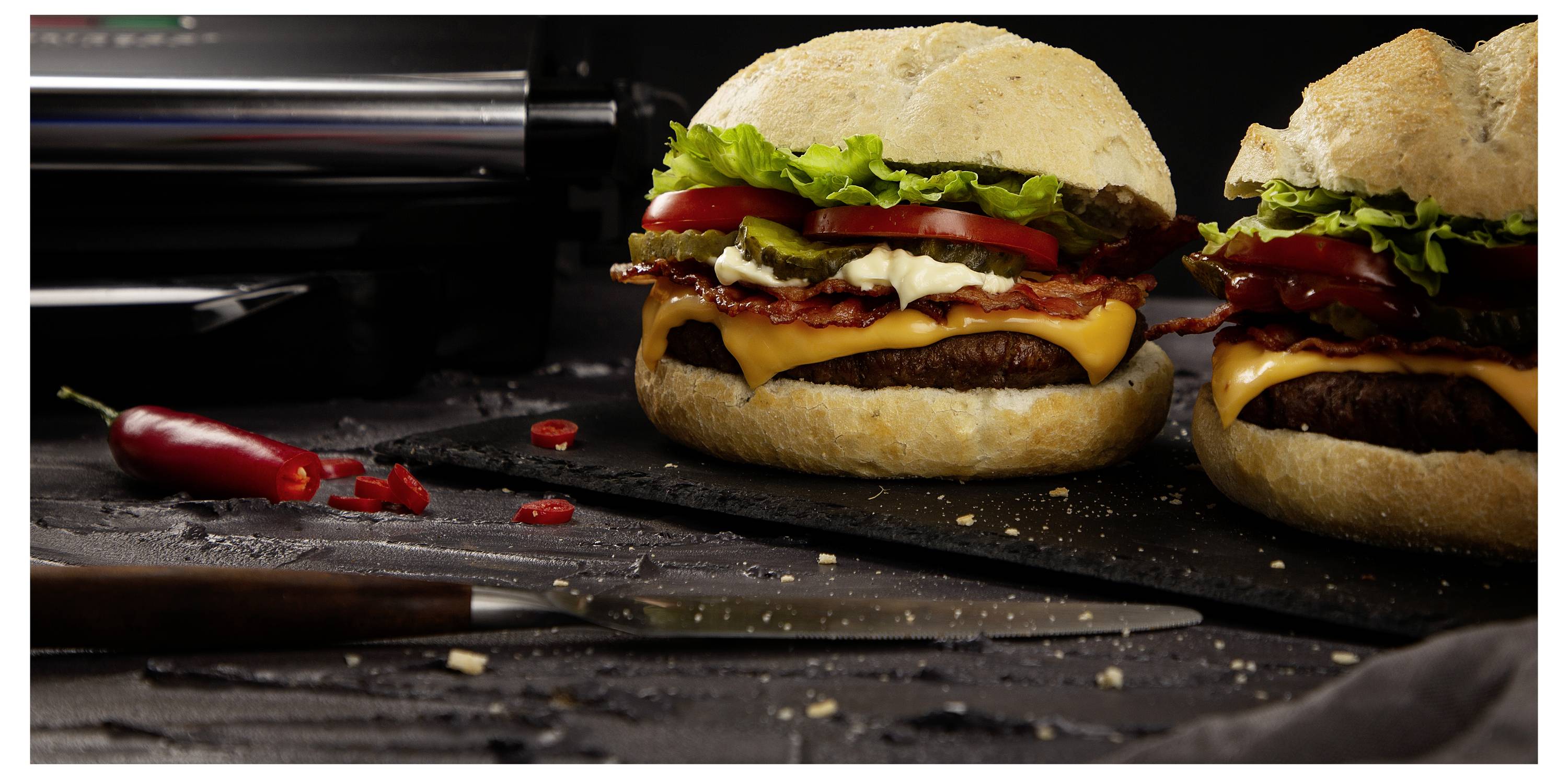 Nahaufnahme von zwei Cheeseburgers mit Salat und Tomate auf einer dunklen Arbeitsfläche, mit einem Messer und einer roten Paprika im Vordergrund.
