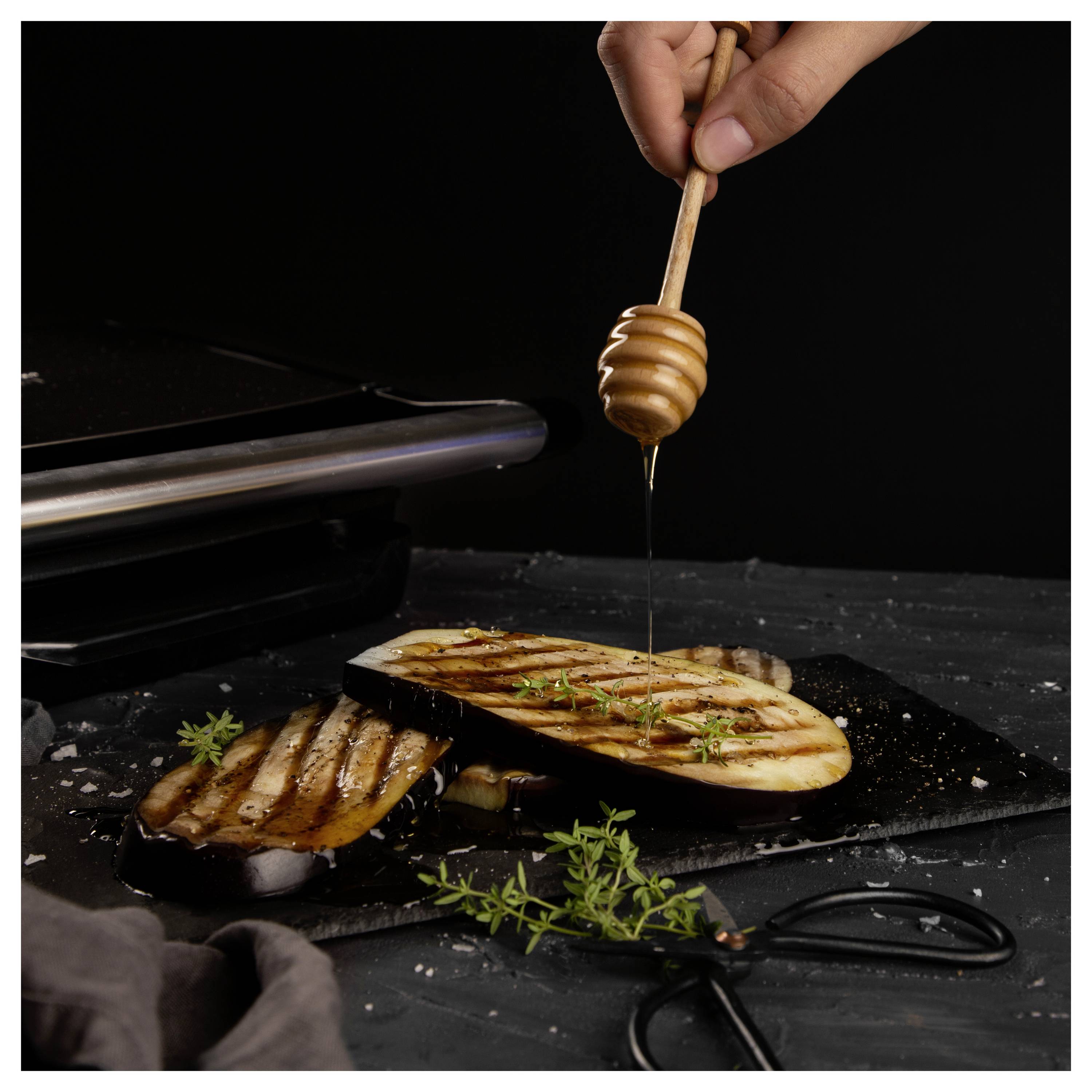 Hand träufelt Honig über gegrilltes Brot oder Käse auf einem dunklen Servierbrett mit Kräutern und einem Honiglöffel.