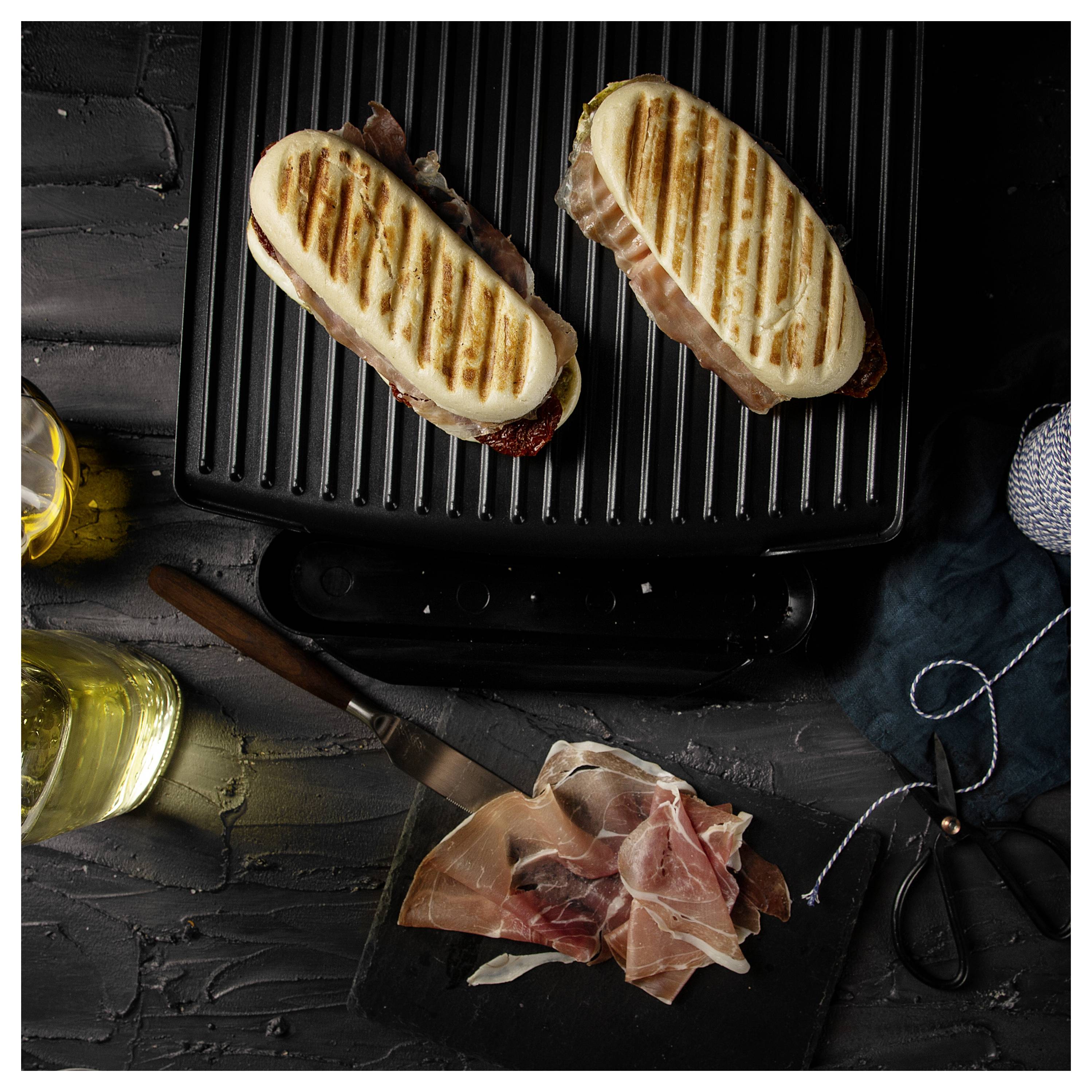 Gegrillte Sandwiches rösten auf einem Kontaktgrill neben geschnittenem Prosciutto, Olivenöl, einem Messer und einer Schere.