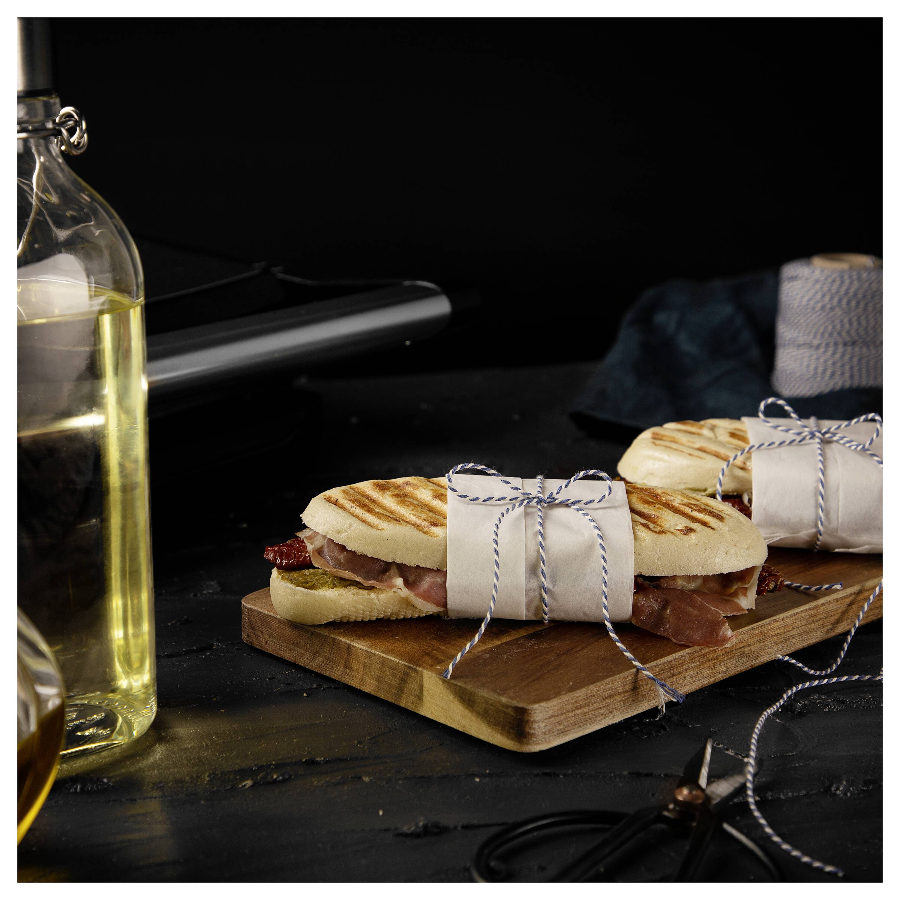 Gegrilltes Sandwich, wie ein Geschenk eingewickelt, auf einem Holzbrett, mit einer Ölflasche daneben auf einer dunklen Tischplatte.