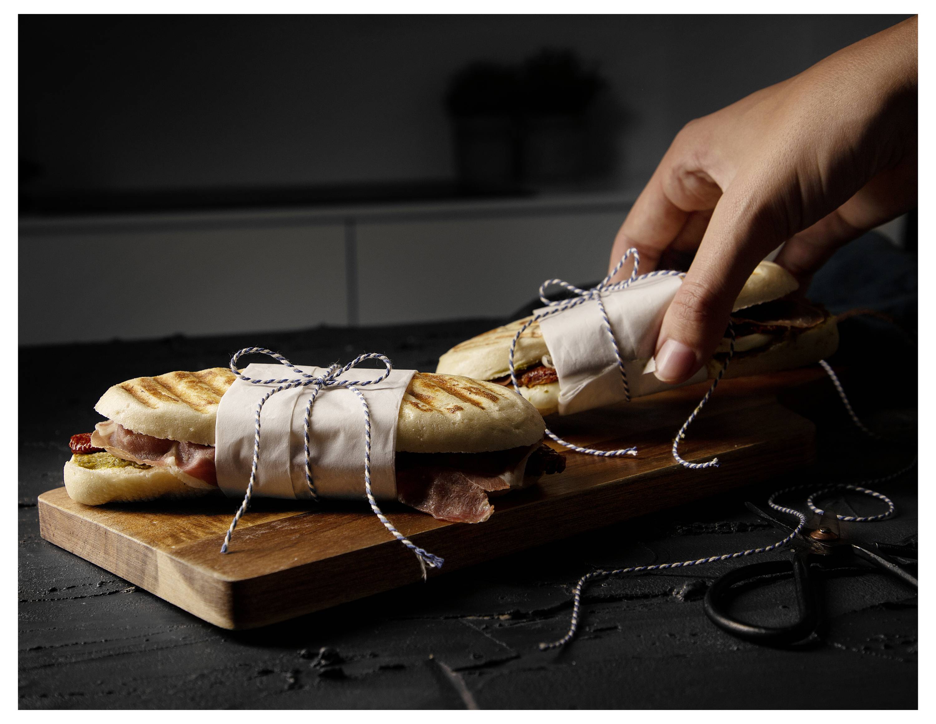 Eine Hand legt ein verpacktes Sandwich auf ein Holzbrett neben einem anderen Sandwich und einer Schere, was zeigt, wie Lebensmittel verpackt oder serviert werden.