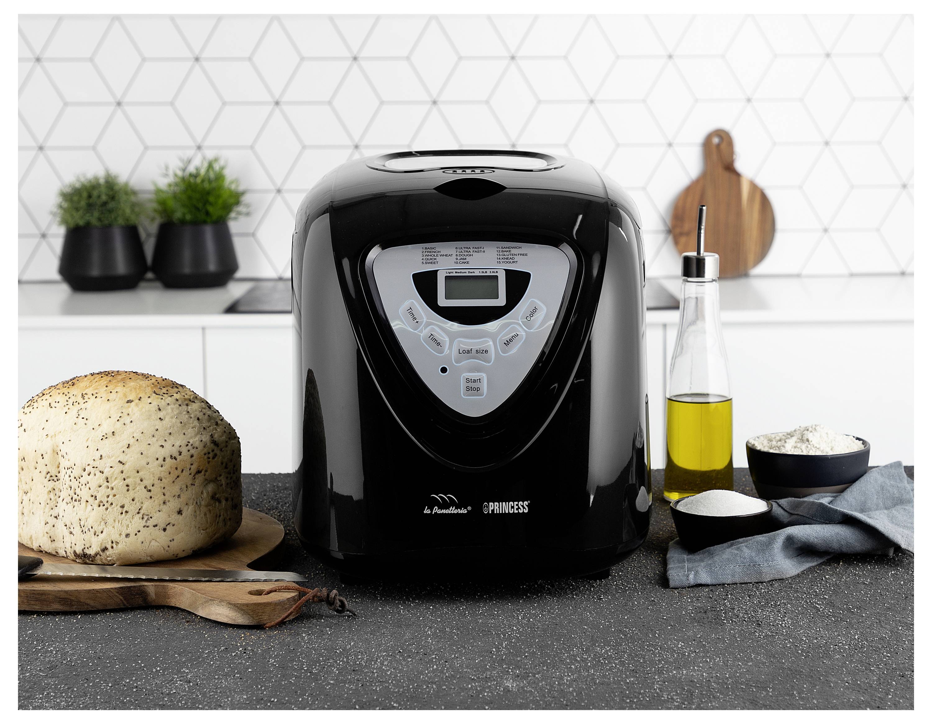 Schwarzbrotbackautomat auf einer Küchenarbeitsfläche, umgeben von einem Laib, Mehl, Öl und Utensilien, was auf die Zubereitung von selbstgebackenem Brot hindeutet.