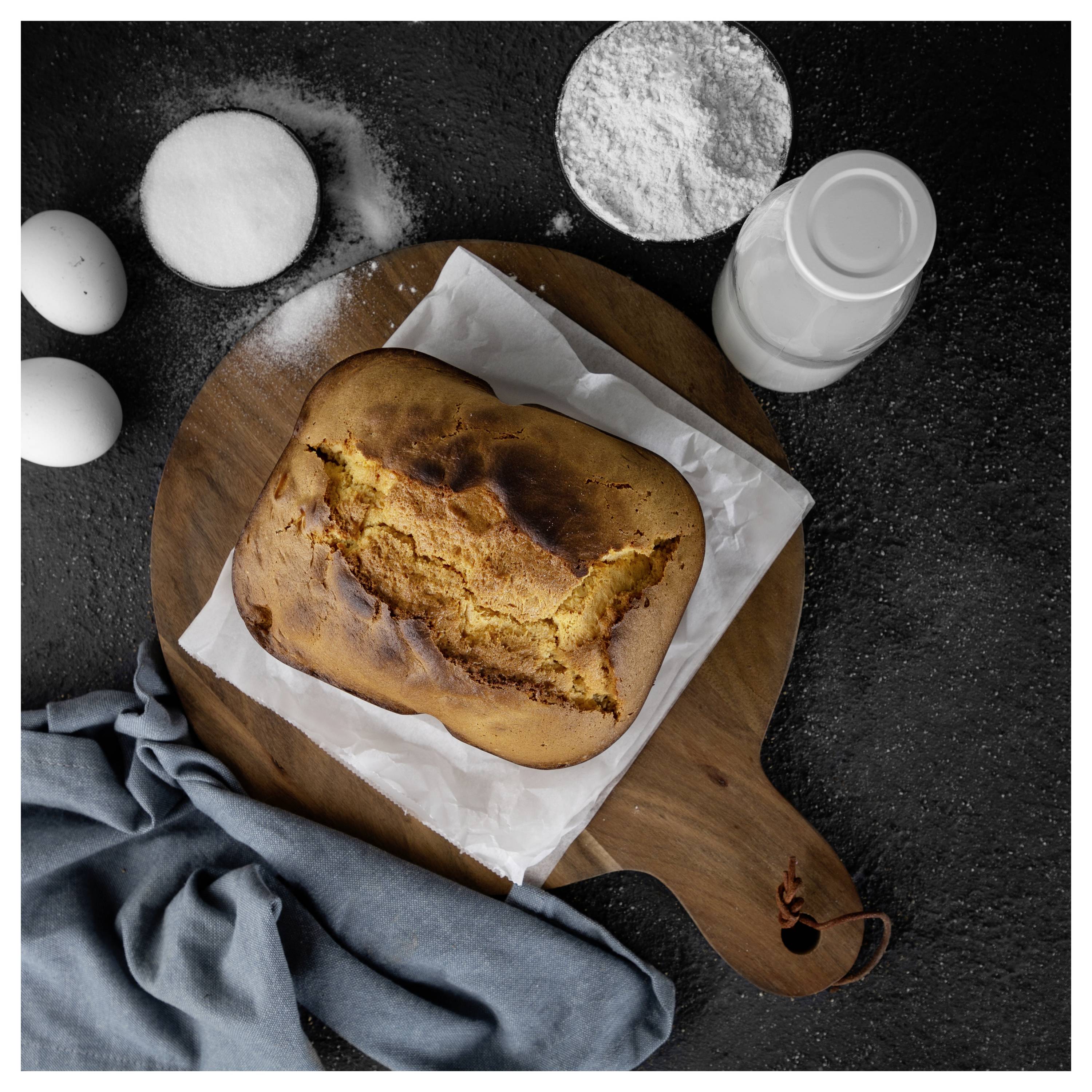 Frisch gebackenes Brot auf Pergamentpapier auf einem Holzbrett, umgeben von Mehl, Eiern, Zucker und einer Flasche Milch auf einer dunklen Arbeitsplatte.