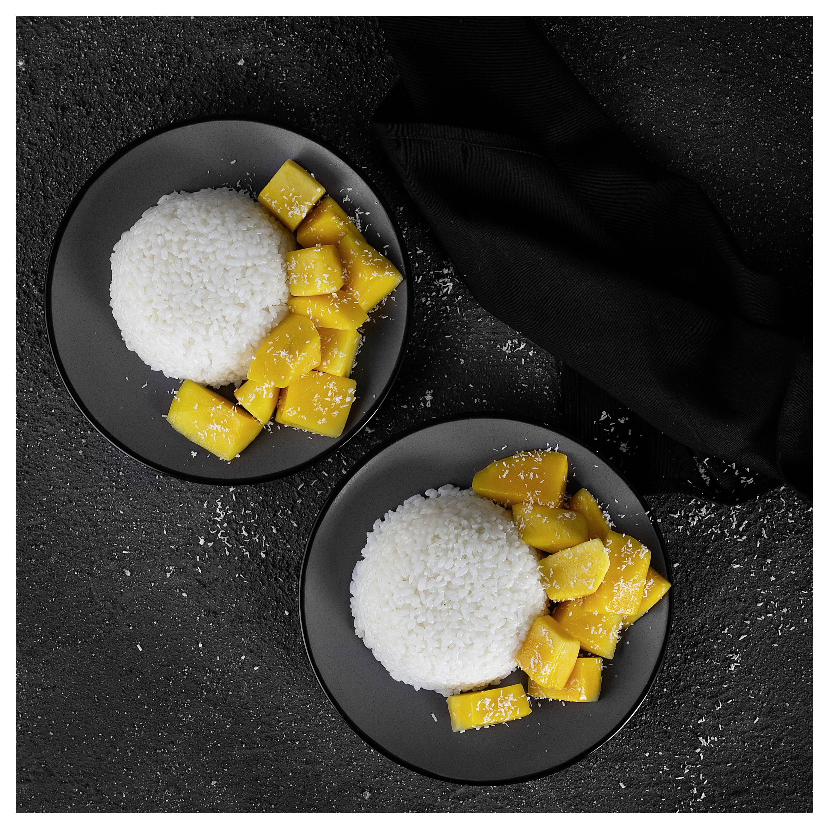 Zwei schwarze Teller halten jeweils einen Haufen weißen klebrigen Reis und geschnittene Mango auf dunklem strukturiertem Hintergrund, mit einer schwarzen Serviette in der Nähe.