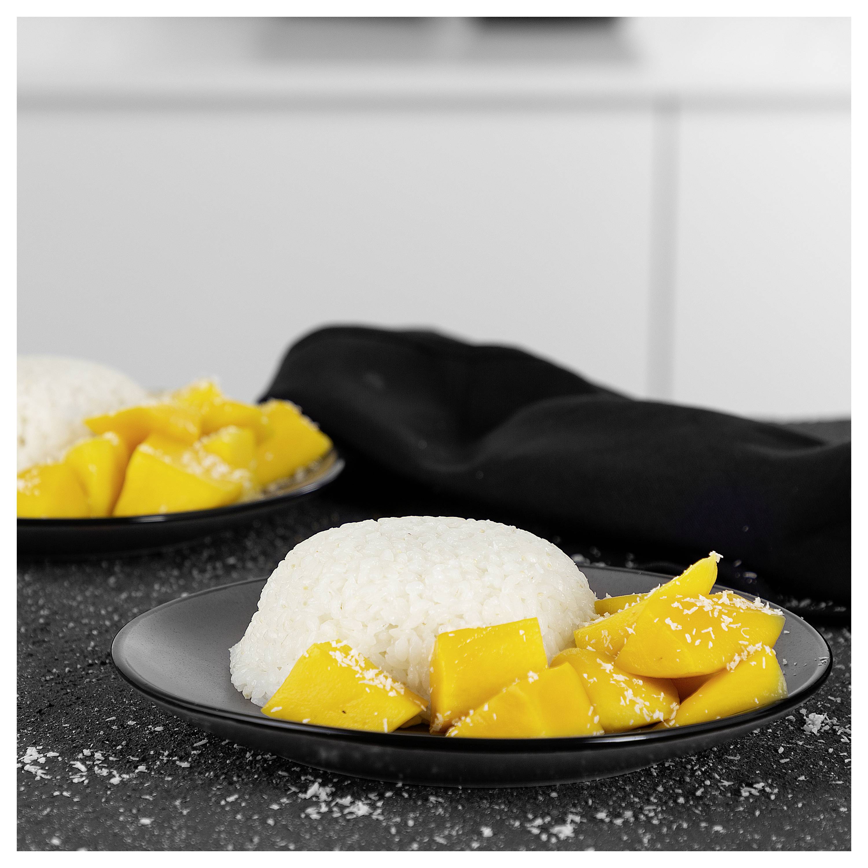 Zwei schwarze Teller mit Kokosnussreis und geschnittener Mango, ein Teller im Vordergrund auf einer Arbeitsplatte.
