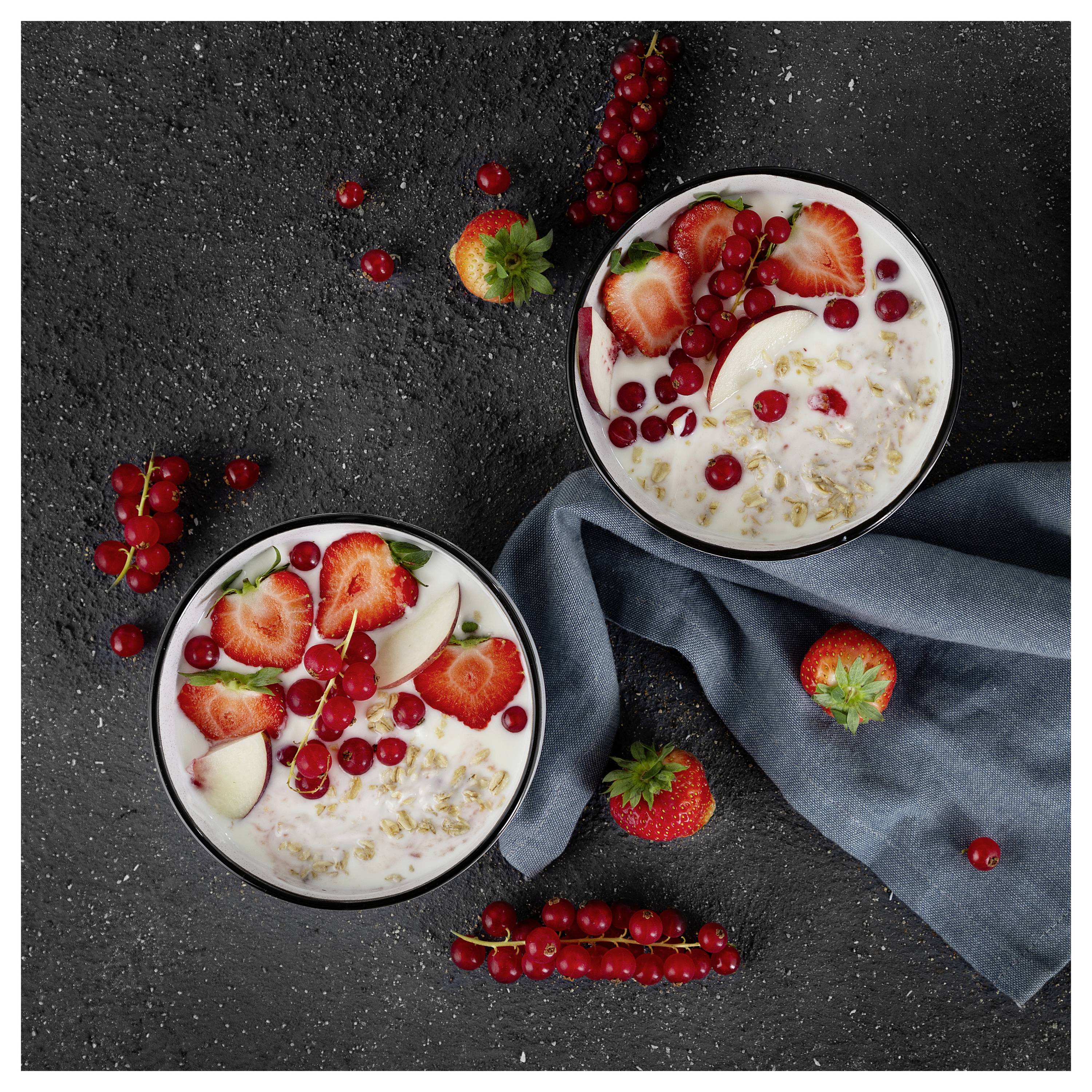 Zwei Schüsseln Joghurt oder Haferflocken, garniert mit Erdbeeren, Apfelscheiben und roten Johannisbeeren auf einer dunklen Oberfläche mit einem blauen Tuch.