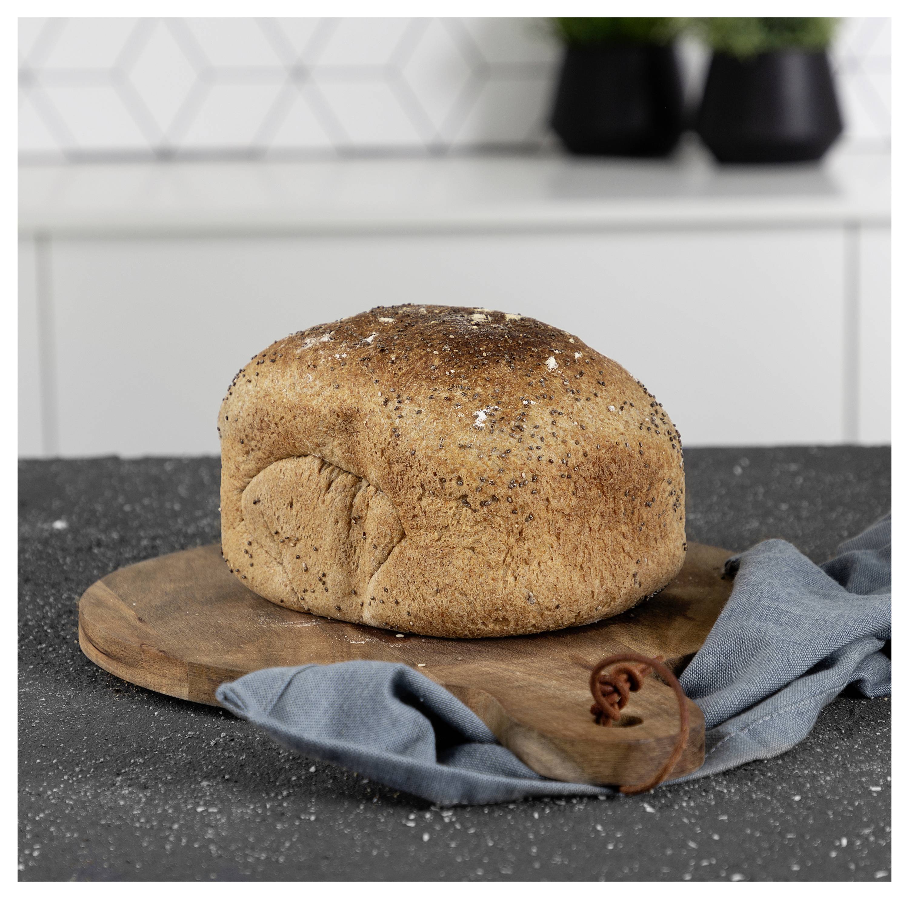 Laib Knusperbrot auf einem Holzbrett mit einem blauen Tuch in einer Küchenszenerie.