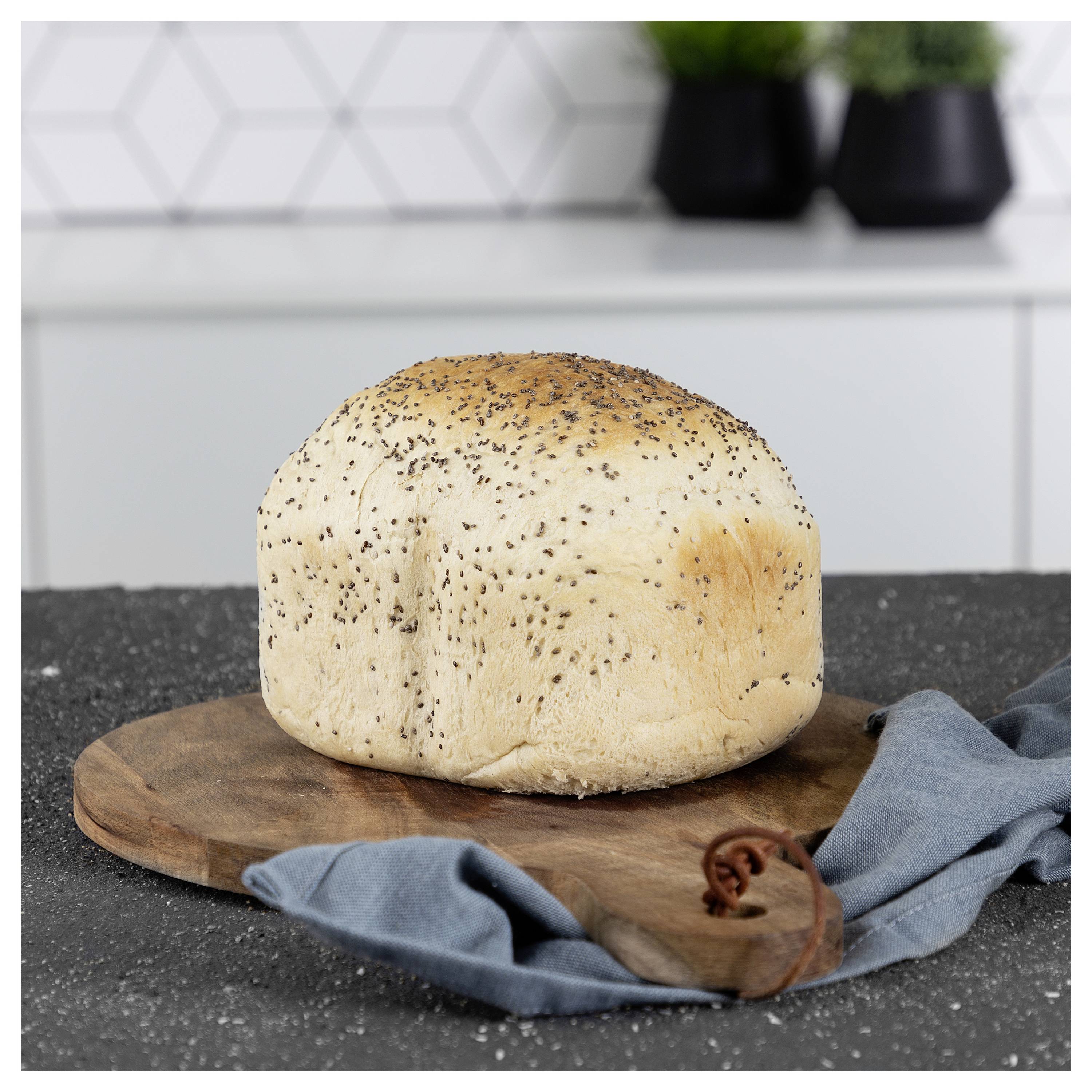Rundes Brotlaib mit Mohnsamen bestäubt auf einem Holzschneidebrett, mit einem blauen Tuch auf einer Küchenarbeitsplatte