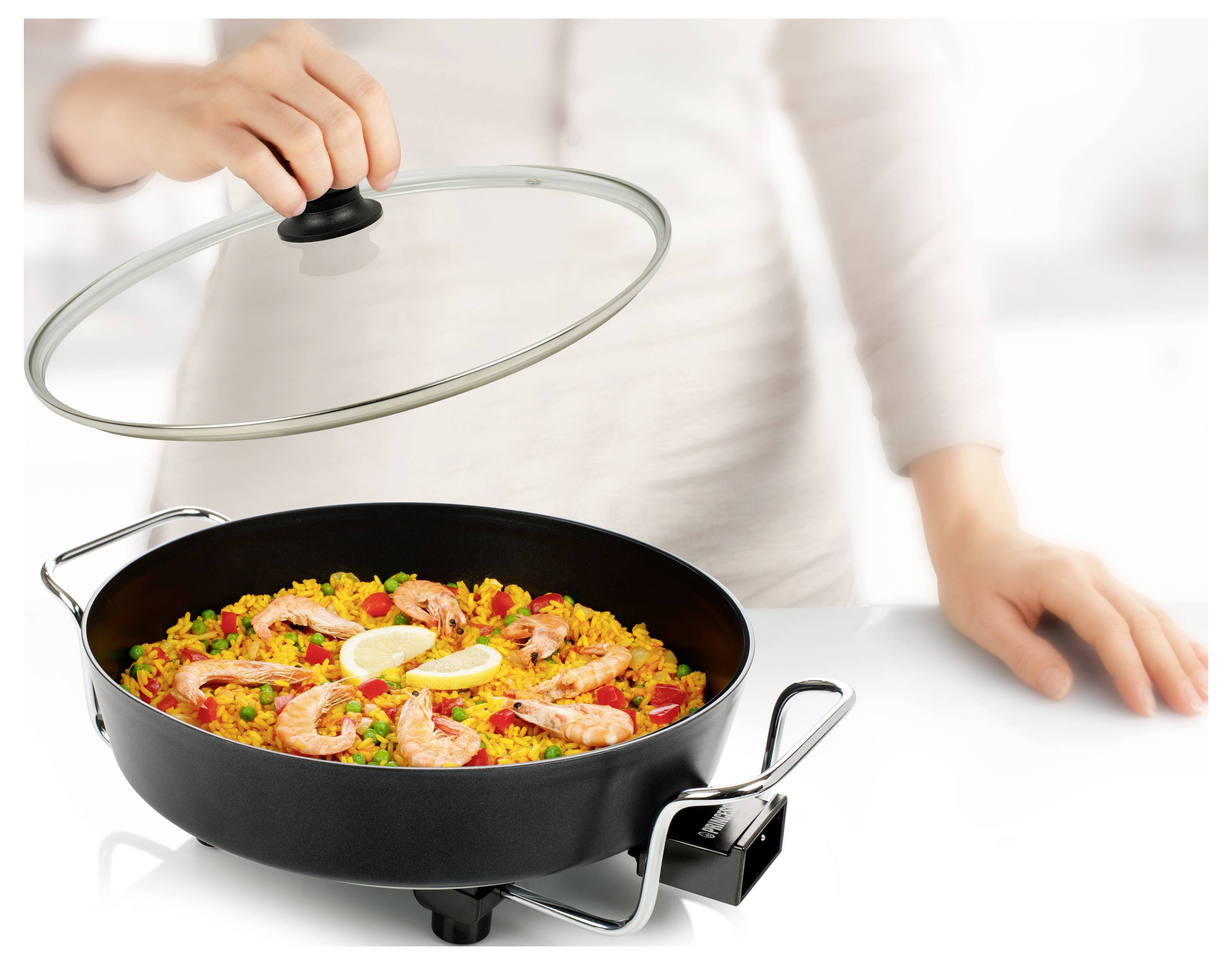 Person, die Meeresfrüchte-Paella in einer großen schwarzen Pfanne zubereitet, hält einen Glasdeckel über dem Gericht auf einem Kochfeld.