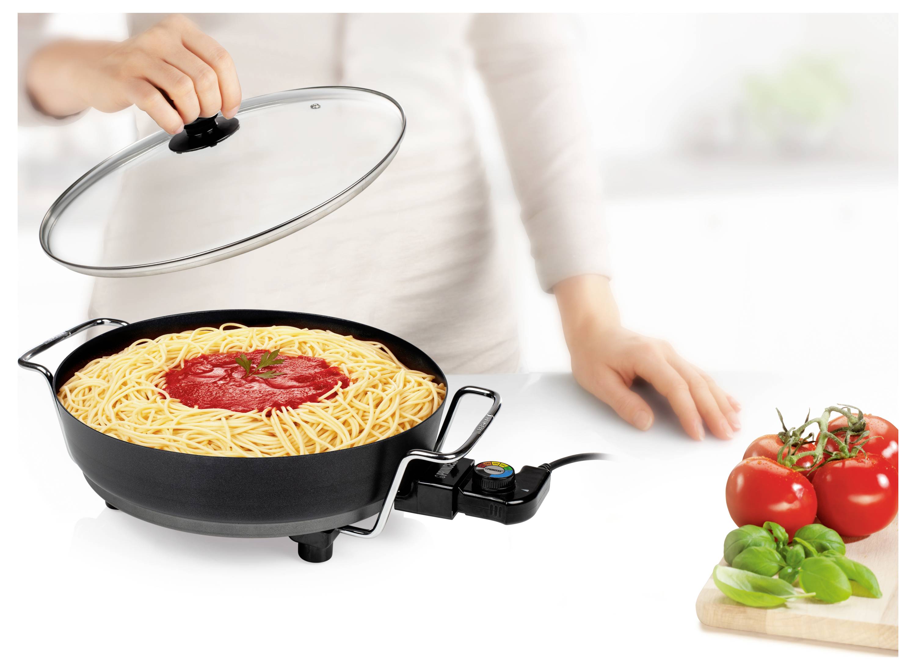 Eine Person hebt einen Deckel an, um Spaghetti mit Tomatensoße zu enthüllen, die in einer elektrischen Pfanne neben frischen Tomaten und Basilikum auf einer weißen Arbeitsplatte kocht.