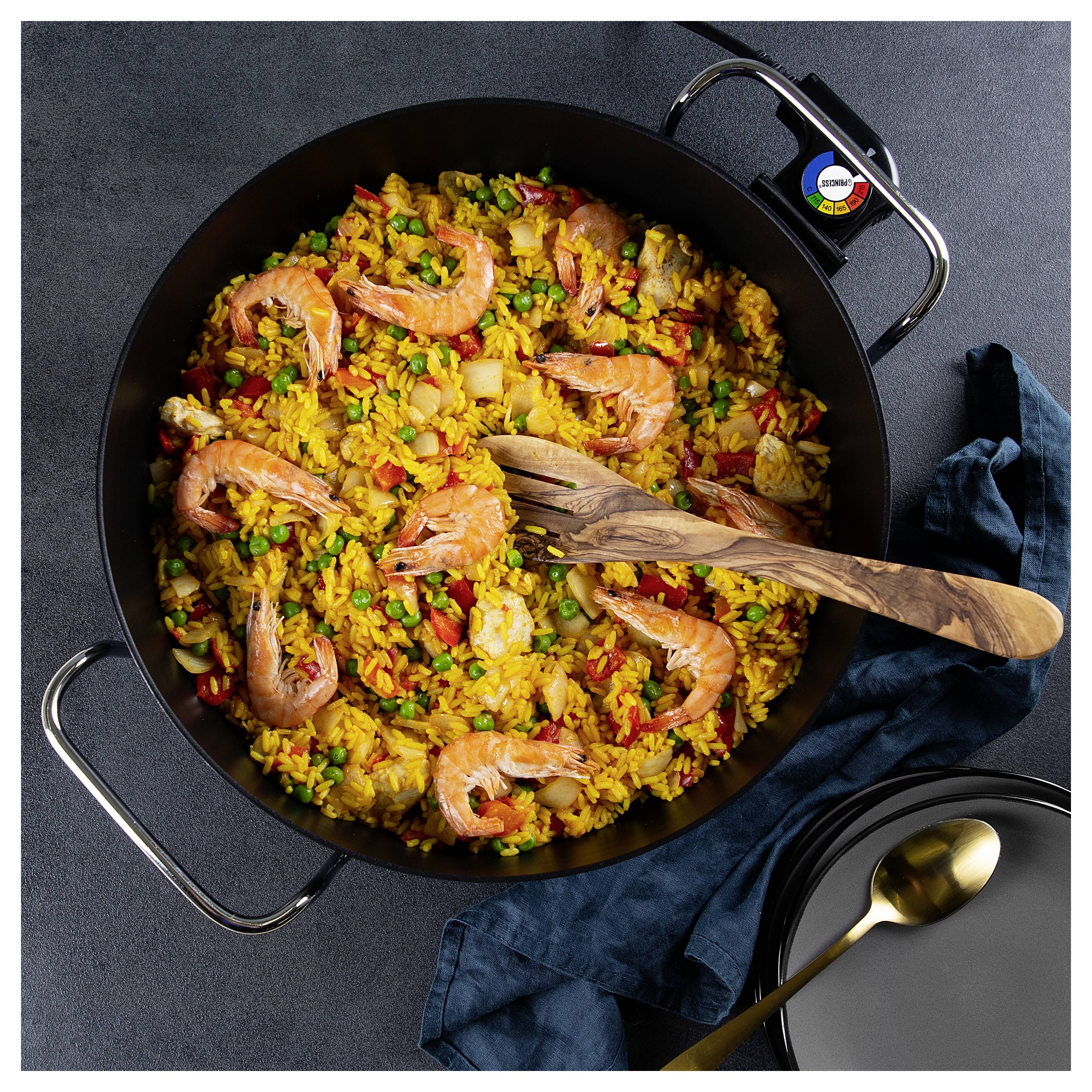 Draufsicht auf eine große Pfanne mit Garnelen-Paella mit Reis, Erbsen, Paprika und einem Holzlöffel auf einem dunklen Tisch.