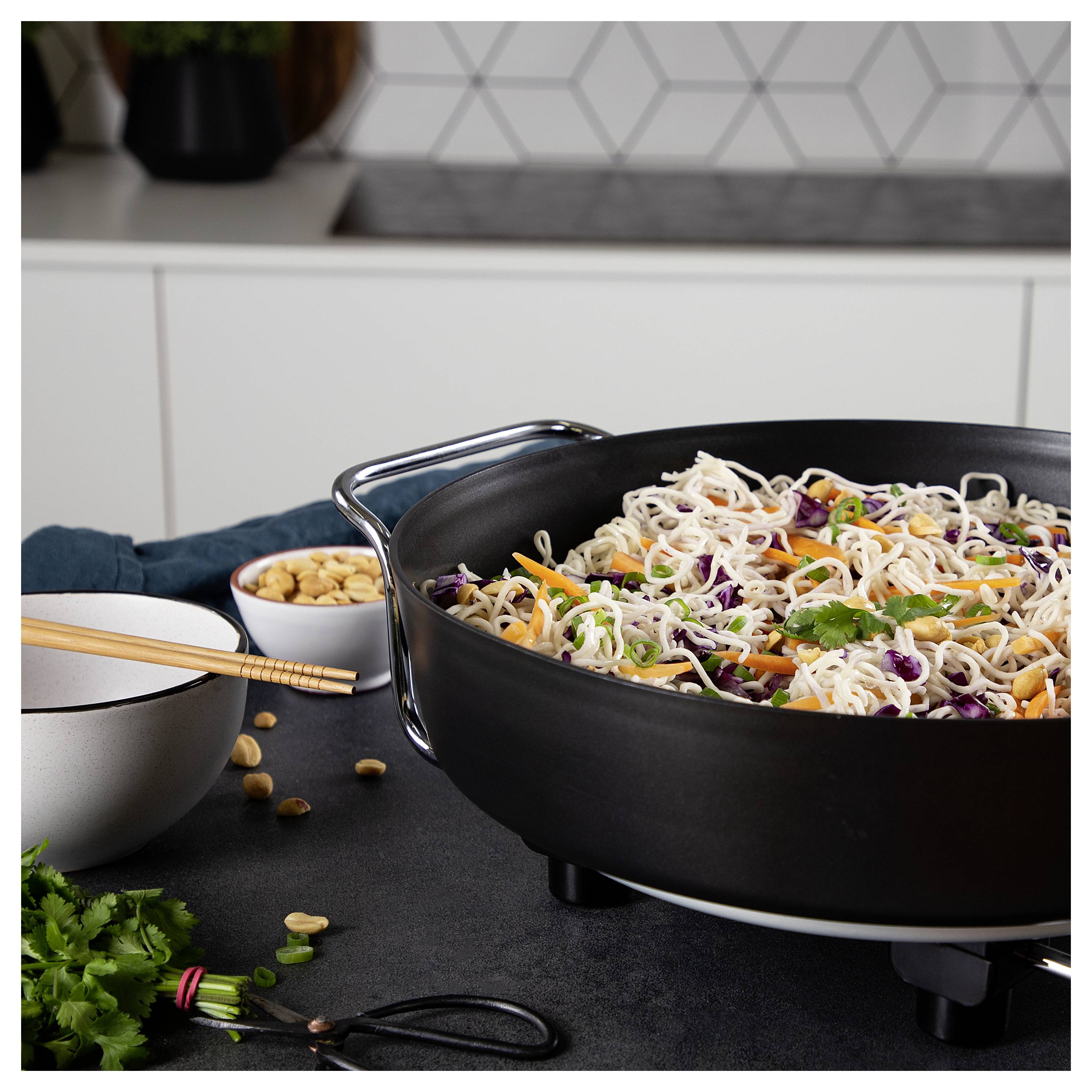 Schwarzer Wok gefüllt mit Nudel-Stir-Fry und Gemüse auf einer Küchentheke, mit Schüsseln, Stäbchen, Erdnüssen und Kräutern in der Nähe.