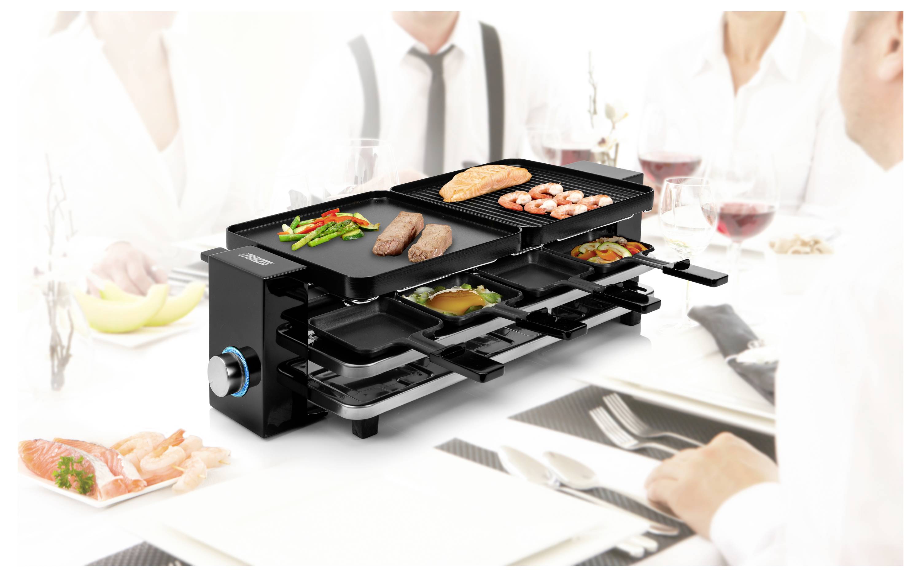Ein tragbarer Elektrogrill mit mehreren Tabletts, auf dem Steaks, Garnelen, Lachs und Gemüse an einem Esstisch gekocht werden, um den herum Menschen sitzen.