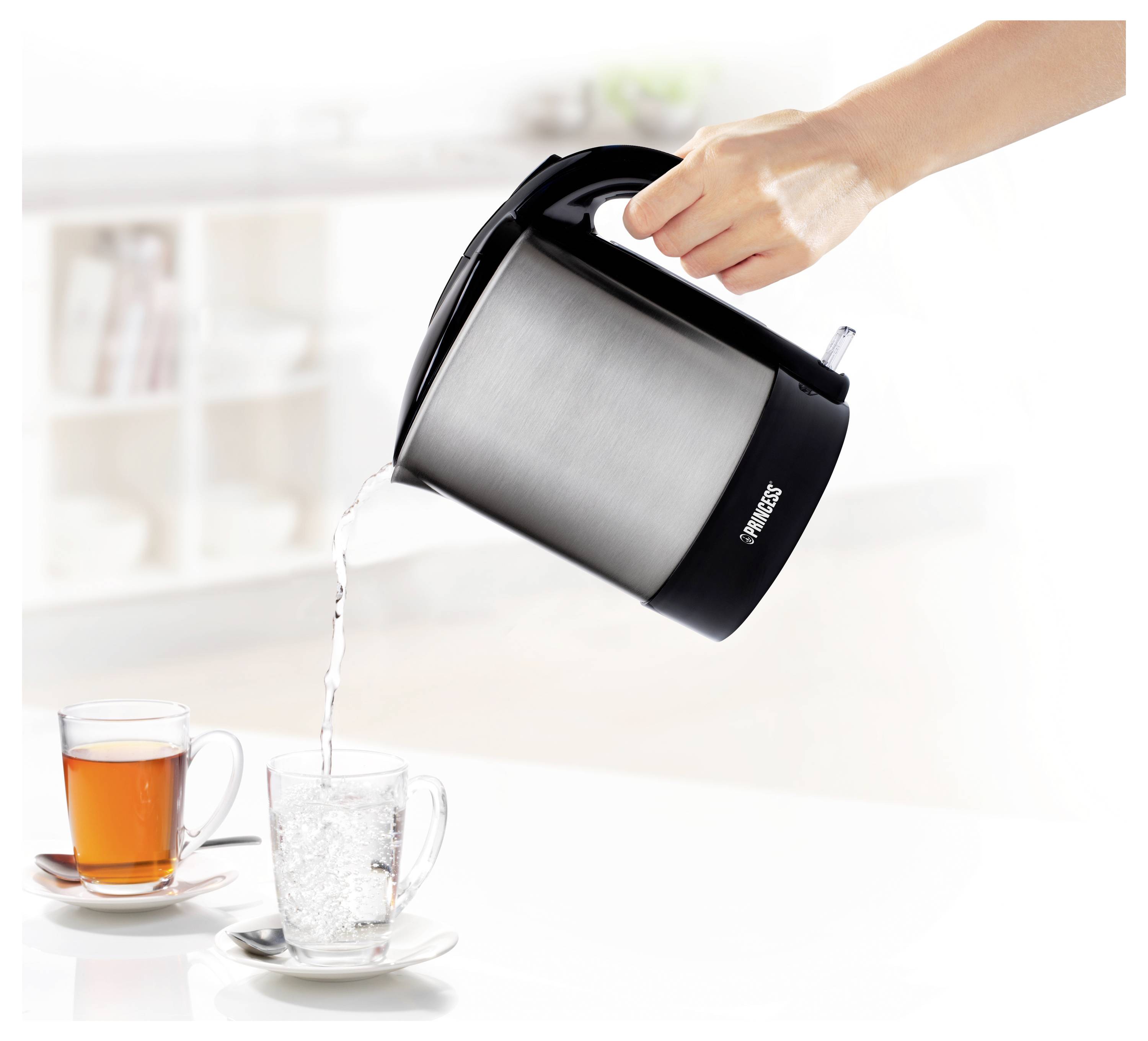 Eine Hand gießt heißes Wasser aus einem Elektrokessel in eine Glastasse neben einer Tasse Tee auf einer hellen Arbeitsplatte.